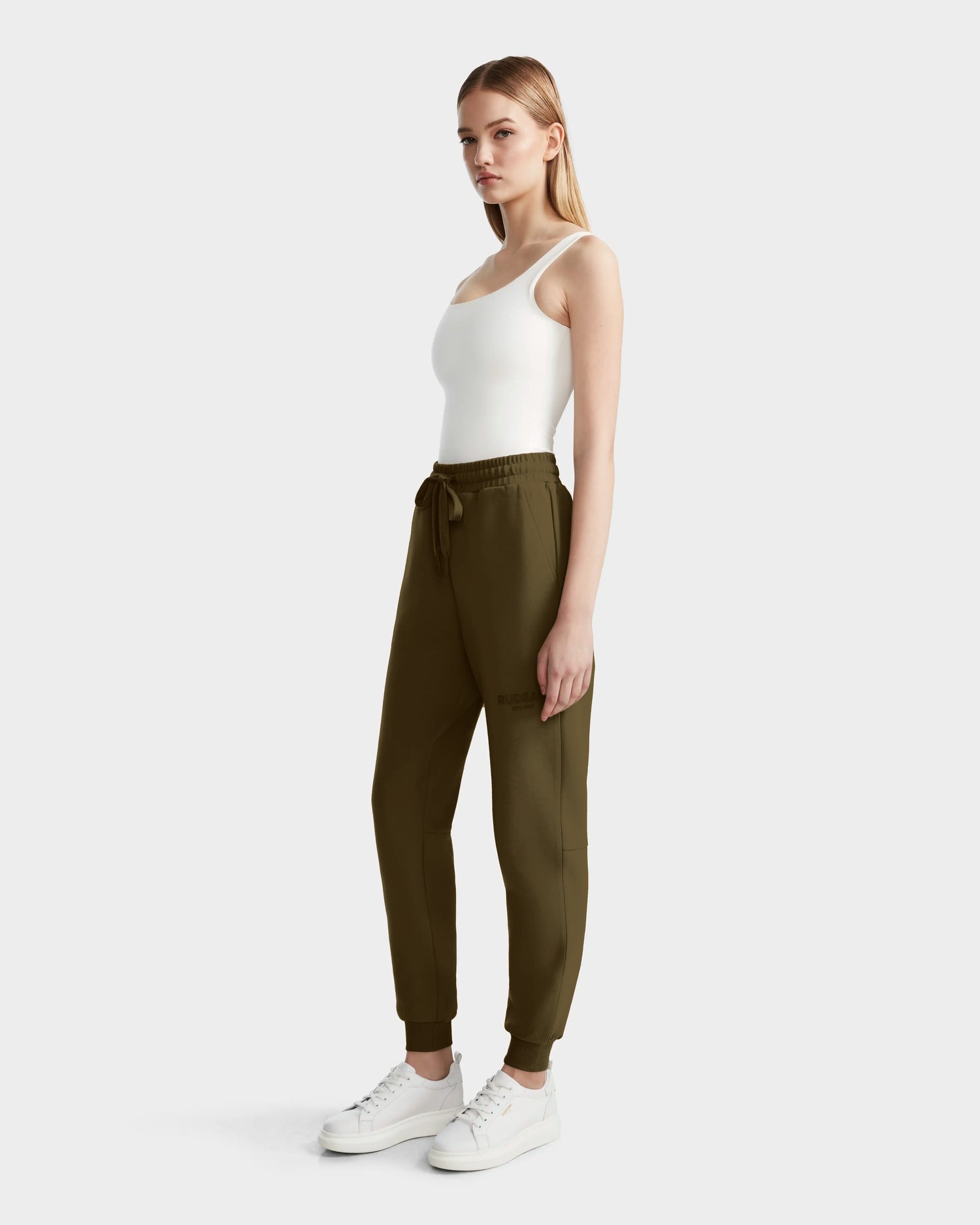 A woman stands against a plain light background in a white tank top, white sneakers, and Rudsak’s JOSS - 9524009 Loden Green W joggers—stylish and crafted from a premium cotton blend with a drawstring waist.