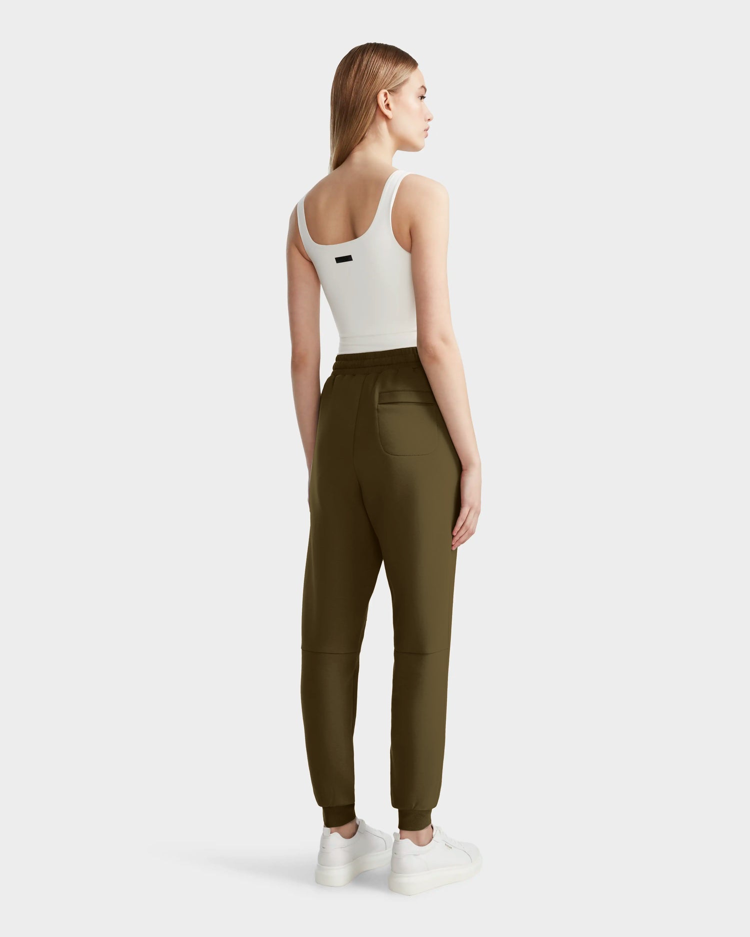 A woman faces away, wearing Rudsak’s JOSS - 9524009 Loden Green W joggers with cuffed ankles and a white crop top. The premium cotton blend pants are styled with white sneakers against a plain light background.