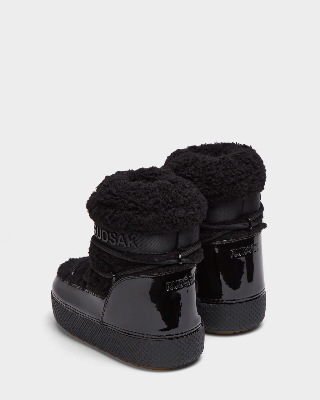 A pair of KATHY - 8223513 Black Rudsak women’s winter boots with shiny patent leather, thick textured soles, and fluffy shearling cuffs, shown from the back against a plain white background.