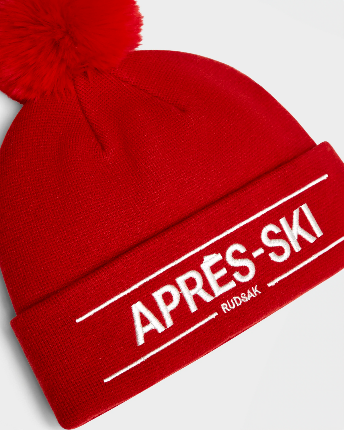 The KRISS - 9424500 Flame Red by Rudsak is a bright red unisex winter hat with a large pom-pom and folded brim, featuring white APRÈS-SKI and RUDSAK embroidery—perfect for chilly après-ski days.