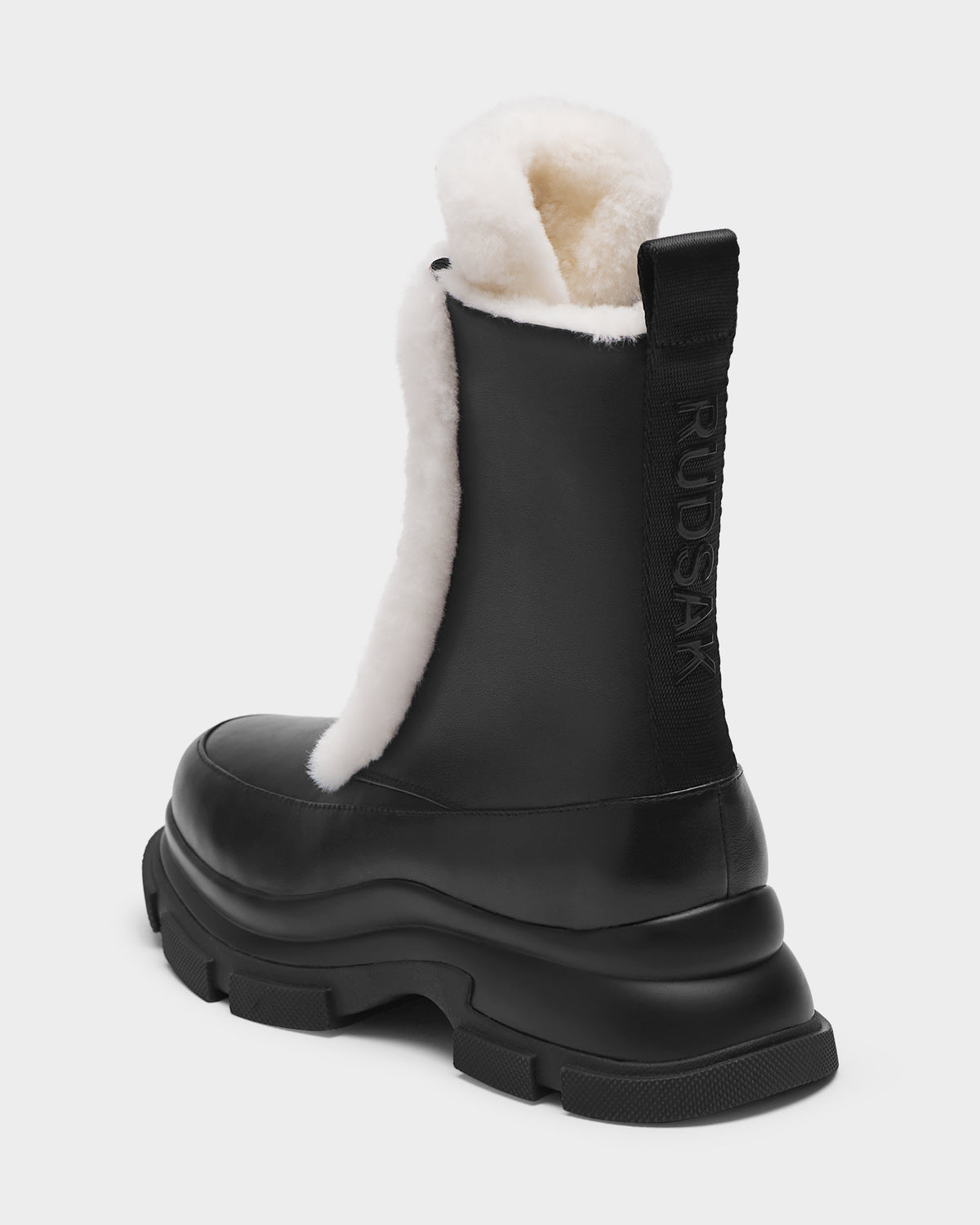The Rudsak LANA S - 8224537 Black is a women’s mid-calf boot in black leather with chunky sole, exposed white shearling lining, and a “RUDAK” embossed pull tab. Shown from the rear side against a white background. Perfect for winter.