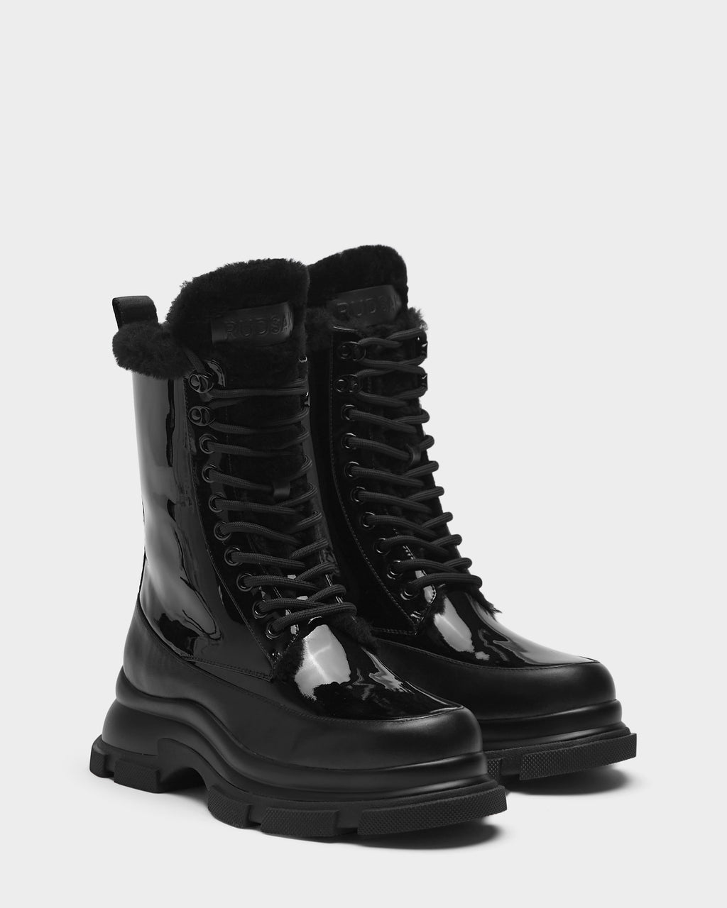 The Rudsak LANA - 8223506 BLACK boots are shiny black mid-calf lace-ups with chunky soles, soft black fur trim at the top, and a cozy shearling lining, shown against a plain white background.