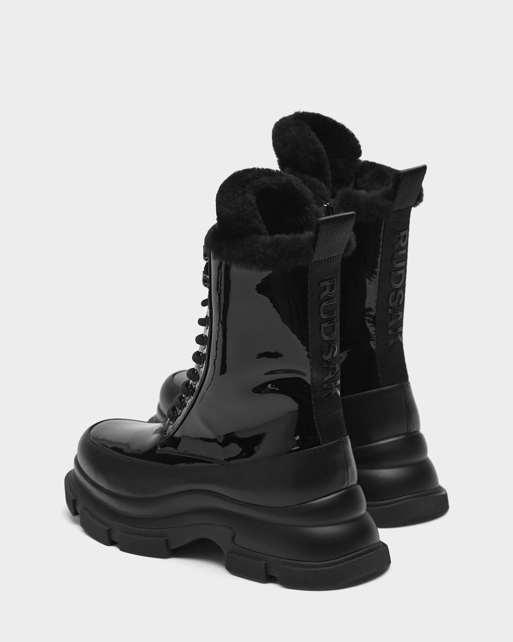 The LANA - 8223506 BLACK boots by Rudsak feature black glossy leather, chunky soles, and a warm shearling lining. RUUXSAX is printed vertically on the heel tabs. Both boots are shown on a plain light background.