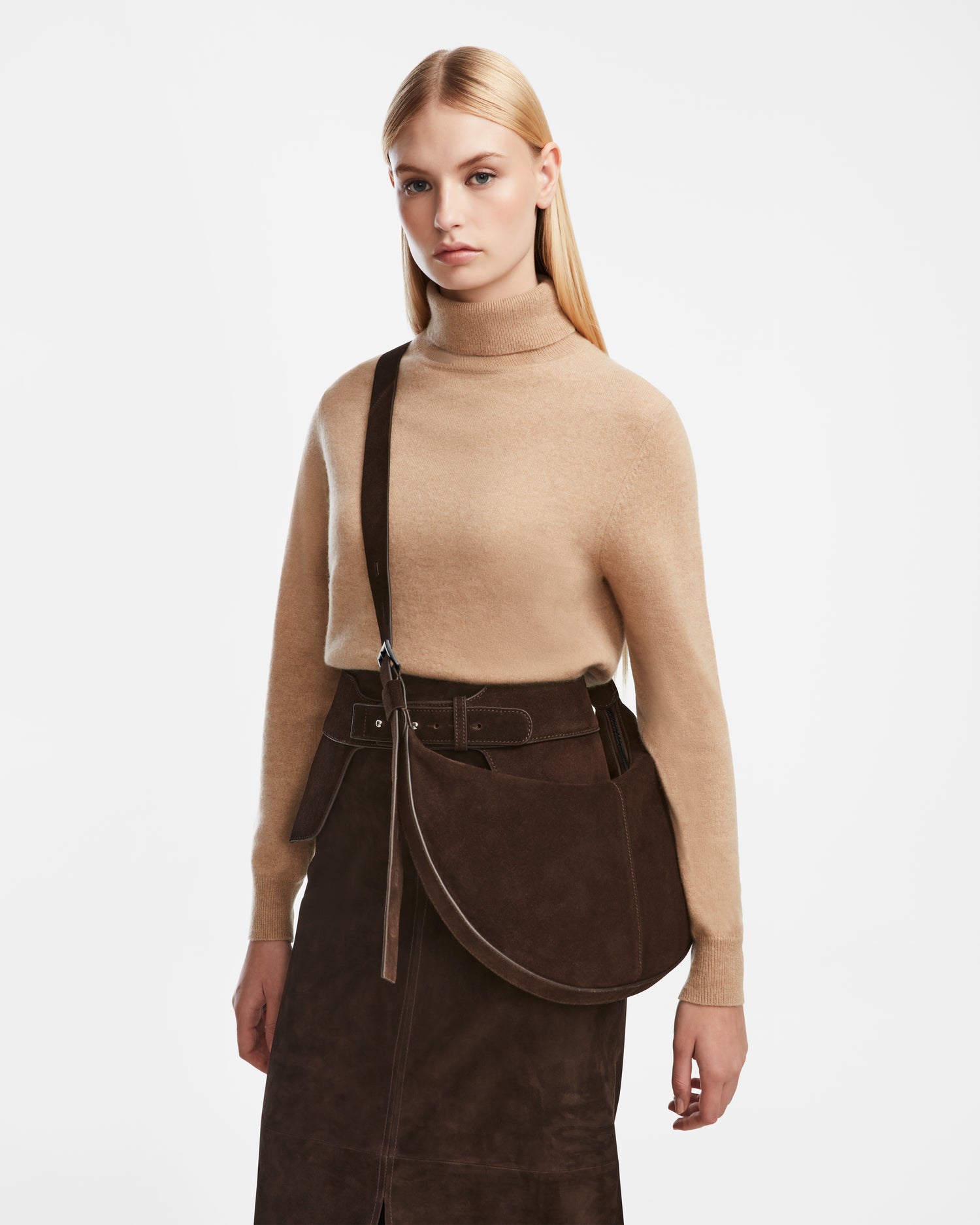 A woman with long blonde hair stands against a white background, wearing a beige turtleneck, dark brown suede skirt, and carrying the Rudsak LANISSA S - 8324520 Espresso suede hobo bag.