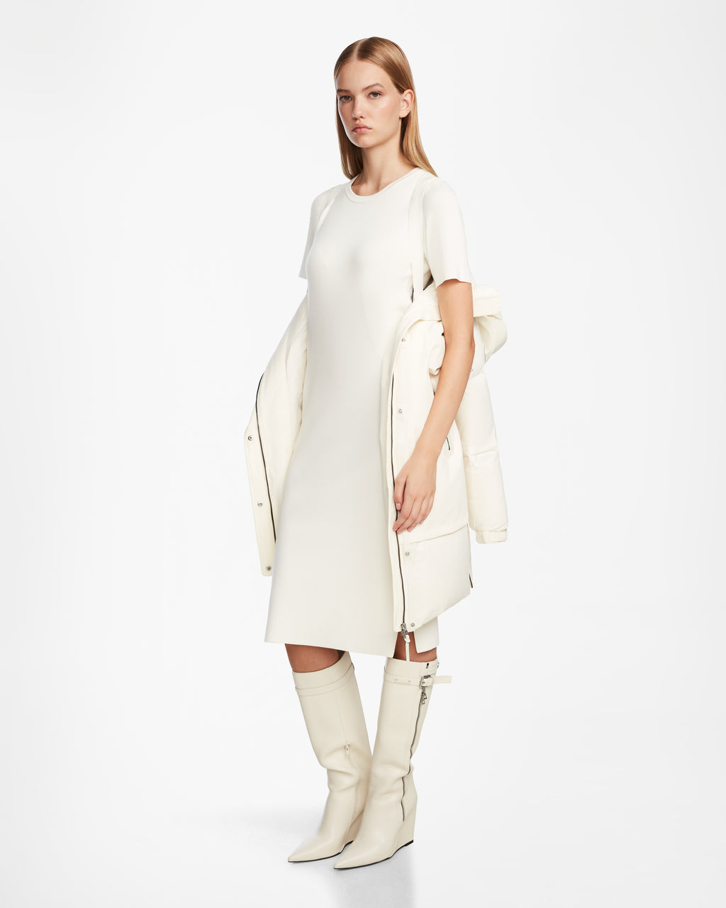 A woman stands against a plain white background wearing the Rudsak LAYAN - 8124576 Iceland women’s down parka draped over her shoulders, paired with a knee-length, short-sleeve white dress and knee-high white boots.