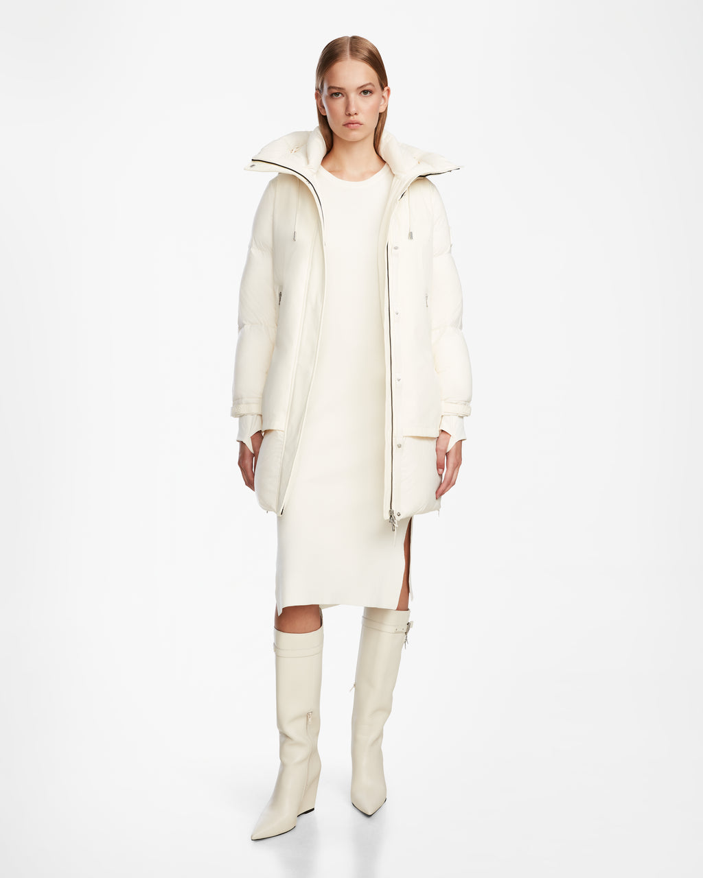 A woman stands against a white background wearing the Rudsak LAYAN - 8124576 Iceland long white down parka over a knee-length dress and knee-high boots, looking directly at the camera with a neutral expression.