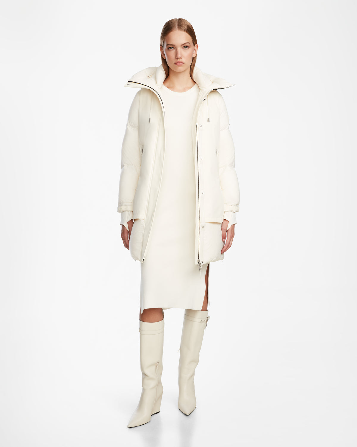 A woman stands against a white background wearing the Rudsak LAYAN - 8124576 Iceland long white down parka over a knee-length dress and knee-high boots, looking directly at the camera with a neutral expression.