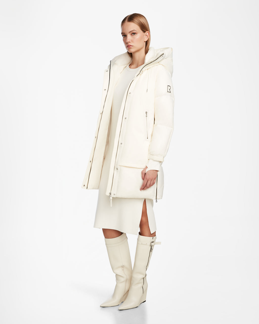 A woman models the Rudsak LAYAN - 8124576 Iceland, a long white winter coat with a hood, over a white knee-length side-slit dress and knee-high white boots, standing against a plain white background.