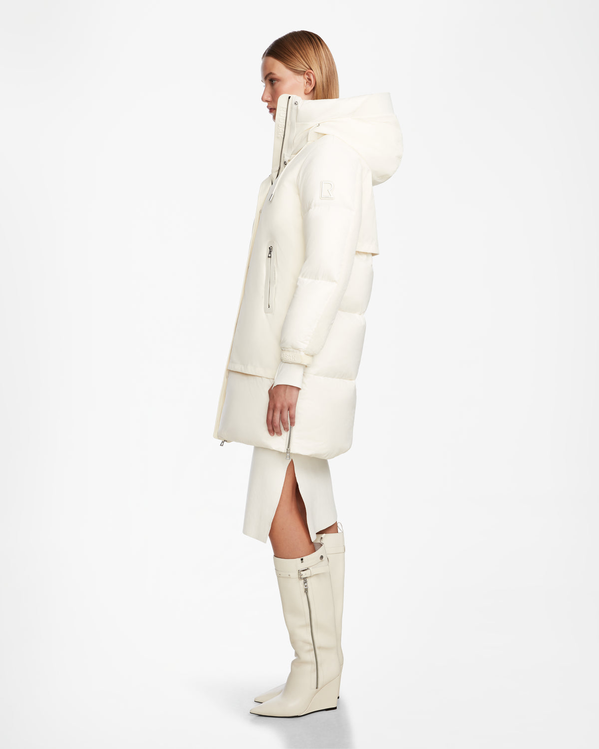 A woman poses in profile against a white background, wearing the Rudsak LAYAN - 8124576 Iceland long white hooded winter coat, paired with white knee-high boots and a front-slit skirt.