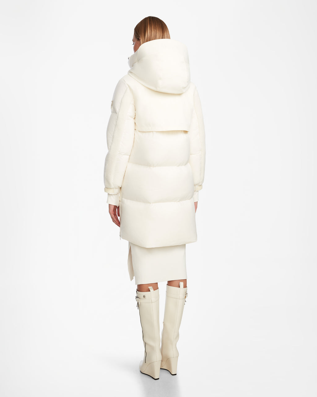 A person with short brown hair stands facing away, wearing the Rudsak LAYAN - 8124576 Iceland long puffy womens down parka with a large hood, paired with a white skirt and knee-high white boots against a plain white background.