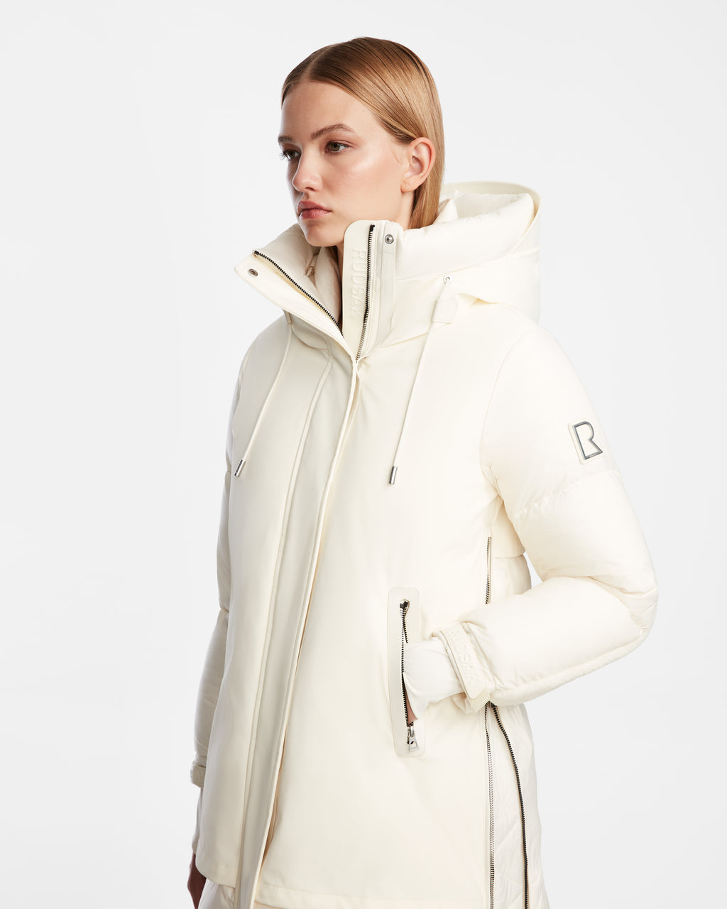 A person with straight, light brown hair wears the Rudsak LAYAN - 8124576 Iceland womens off-white down parka with a high collar, hood, zippered pockets, and an R patch on the sleeve, set against a plain white background.