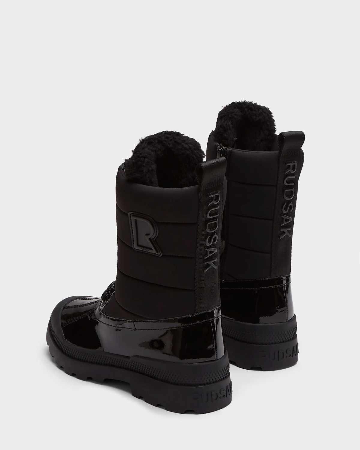 The Rudsak LEX - 8224534 Black women’s winter boots feature shiny patent details, thick soles, faux fur lining, and RUDSAK branding on the back heel and strap. These water-resistant boots are shown on a white background.