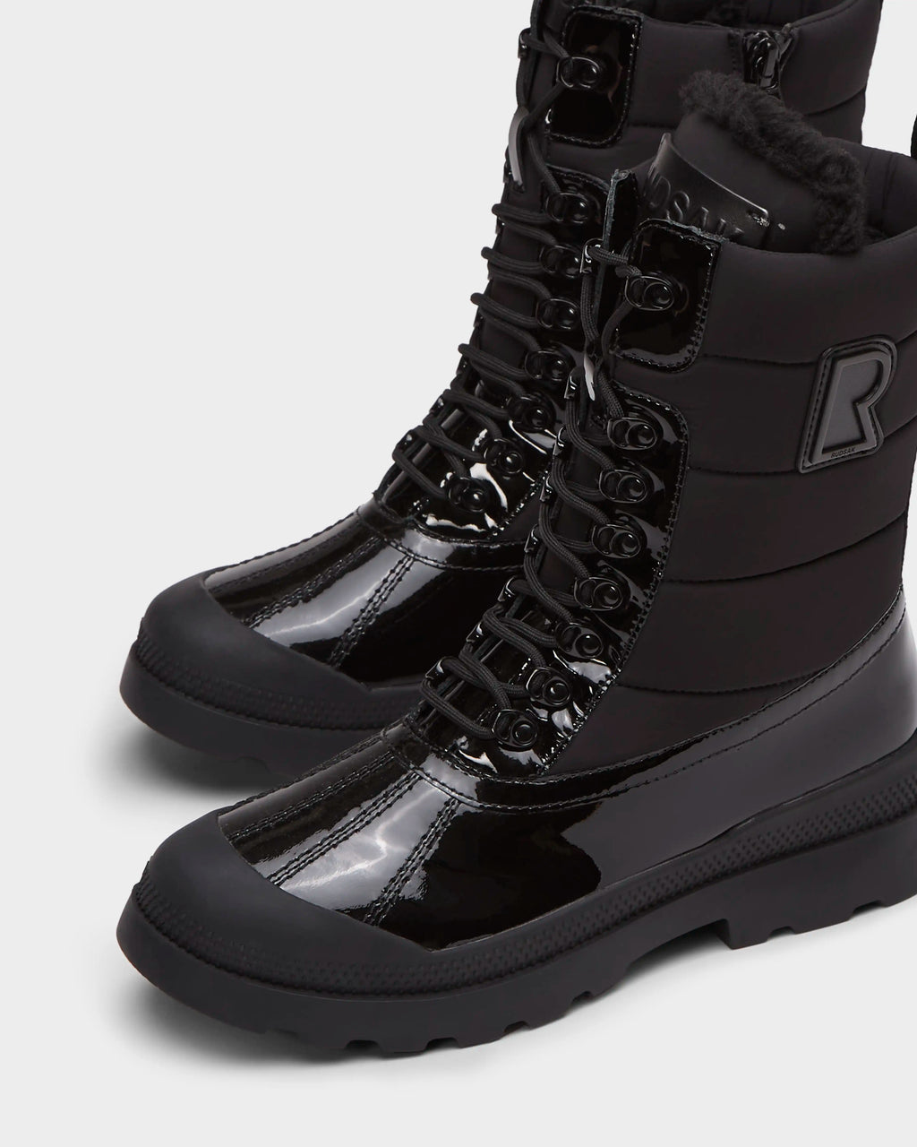 Close-up of LEX - 8224534 Black high-top women’s winter boots by Rudsak, featuring shiny patent leather, thick rubber soles, lace-up fronts, a bold R patch, wool lining for insulation, and a water-resistant design.