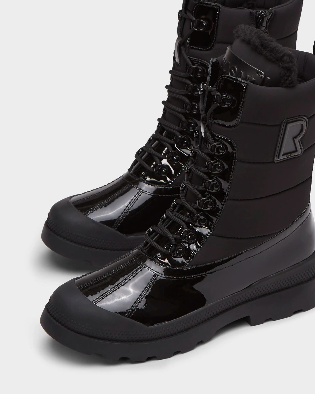Close-up of LEX - 8224534 Black high-top women’s winter boots by Rudsak, featuring shiny patent leather, thick rubber soles, lace-up fronts, a bold R patch, wool lining for insulation, and a water-resistant design.