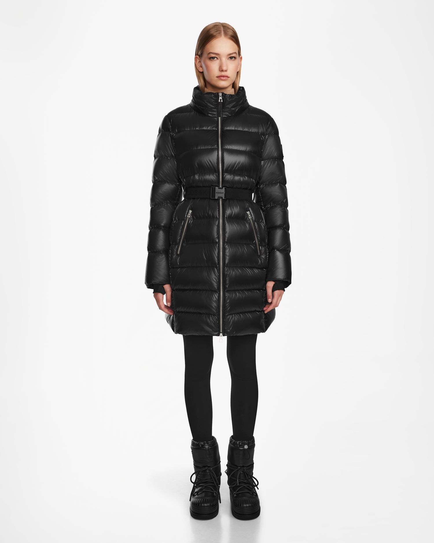 A woman faces forward against a plain white background, wearing the Rudsak LILITH - L124539 Black long quilted winter coat with a belt, paired with black leggings and black boots.