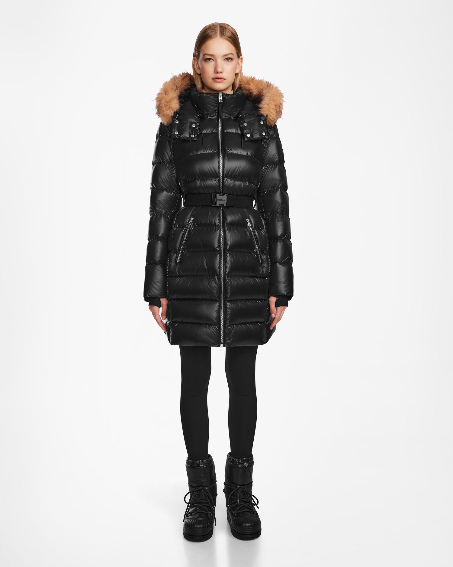 A woman models the Rudsak LILITH - L124539 Black down puffer with a belted waist and fur-trimmed hood, paired with black leggings and boots, standing against a plain white background.