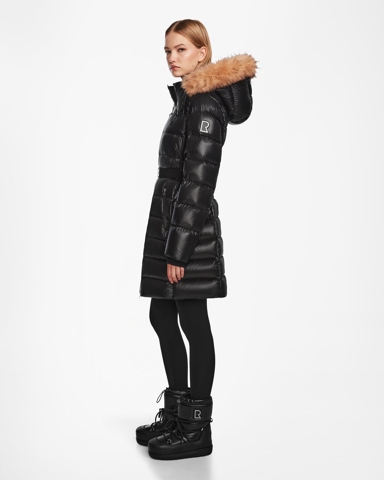 A woman stands sideways wearing the Rudsak LILITH - L124539 Black down puffer with a fur-trimmed hood, paired with black leggings and winter boots, against a plain white background.