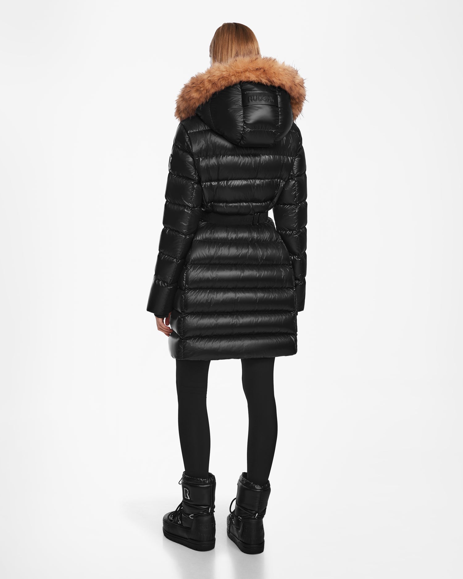 A person faces away from the camera wearing the Rudsak LILITH - L124539 Black women's down puffer with a brown fur-trimmed hood, paired with black leggings and winter boots, set against a white background.