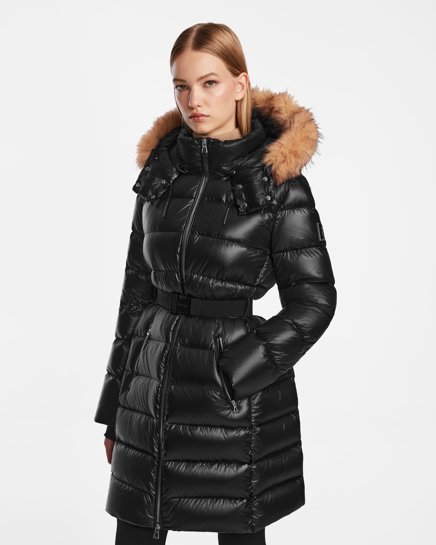 A woman wears the Rudsak LILITH - L124539 Black quilted winter coat with a fur-trimmed hood and belted waist, standing against a plain white background.