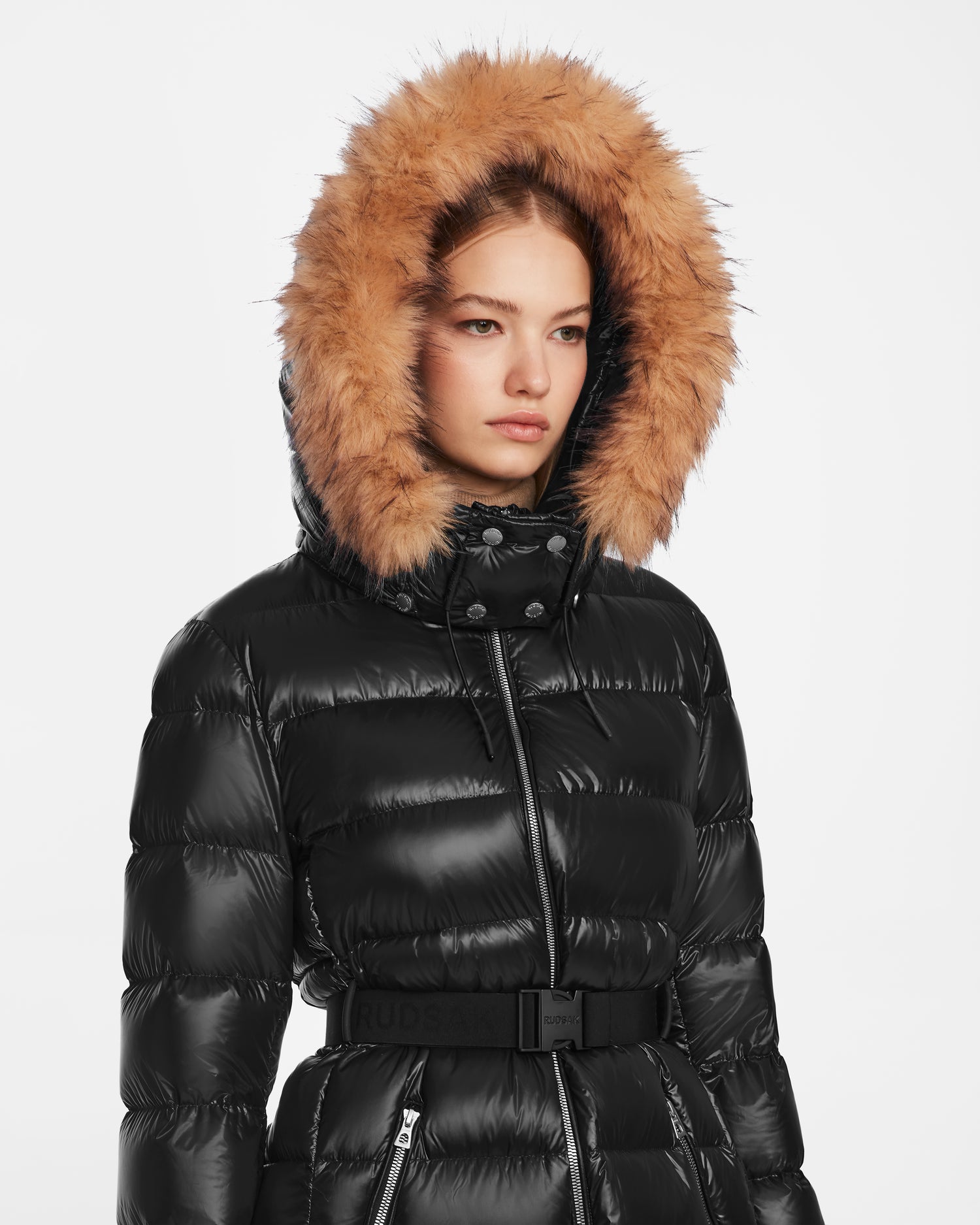 A woman wears the Rudsak LILITH - L124539 Black quilted winter coat with a fur-lined hood and light brown trim, standing facing forward against a plain white background.