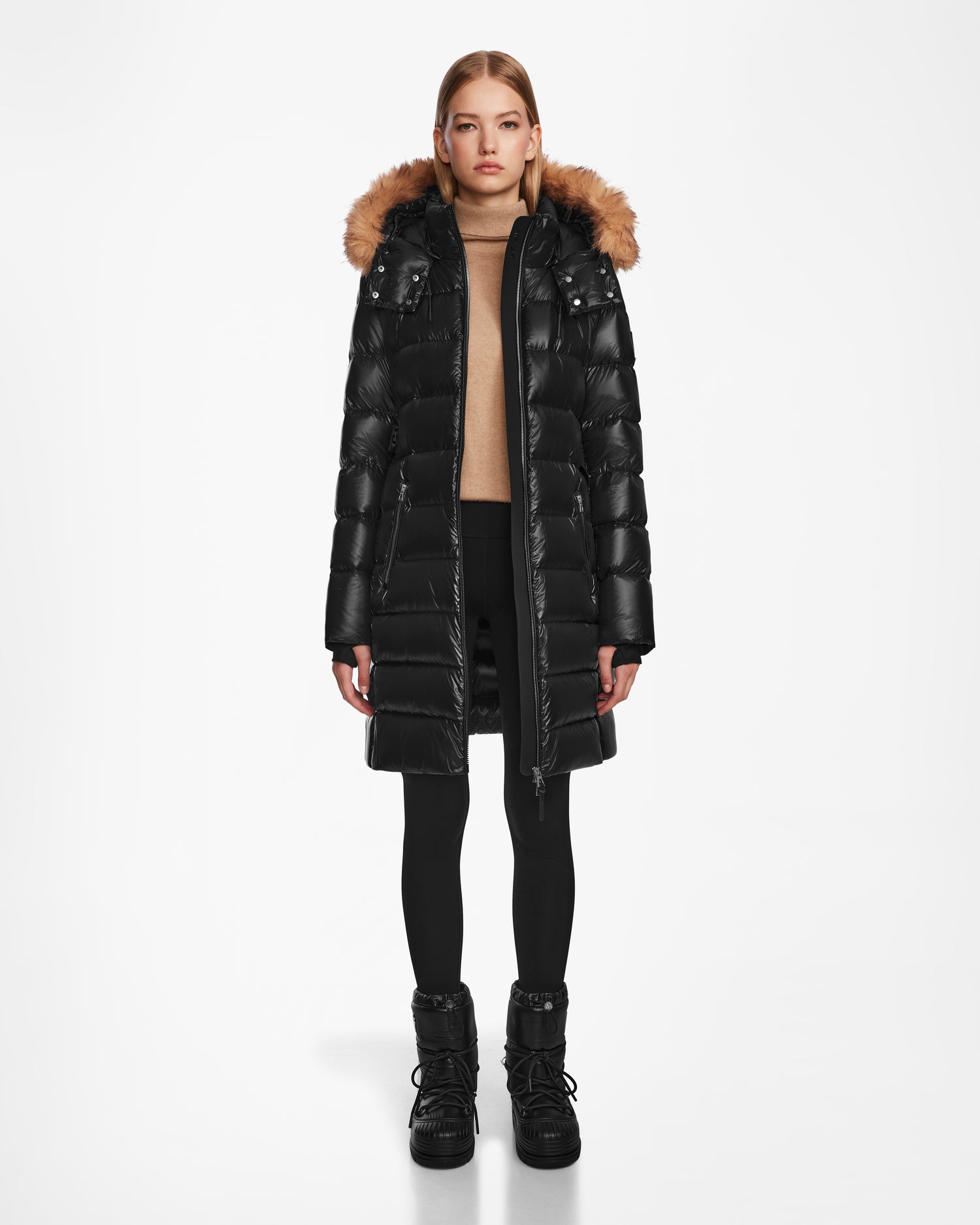 A woman faces forward against a white background, wearing the Rudsak LILITH - L124539 Black long quilted winter coat with a fur-trimmed hood, paired with black leggings, black boots, and a beige turtleneck sweater.