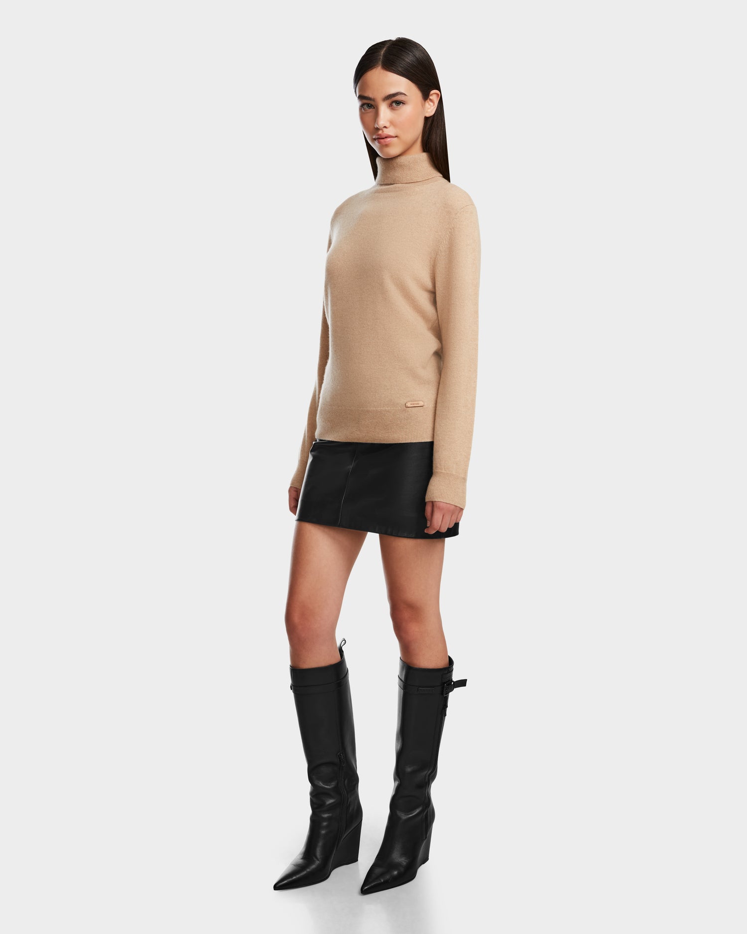 A woman with straight dark hair and a neutral expression models the Rudsak LIZA - 8524598 Camel women’s cashmere turtleneck, paired with a black leather mini skirt and black knee-high heeled boots against a plain background.