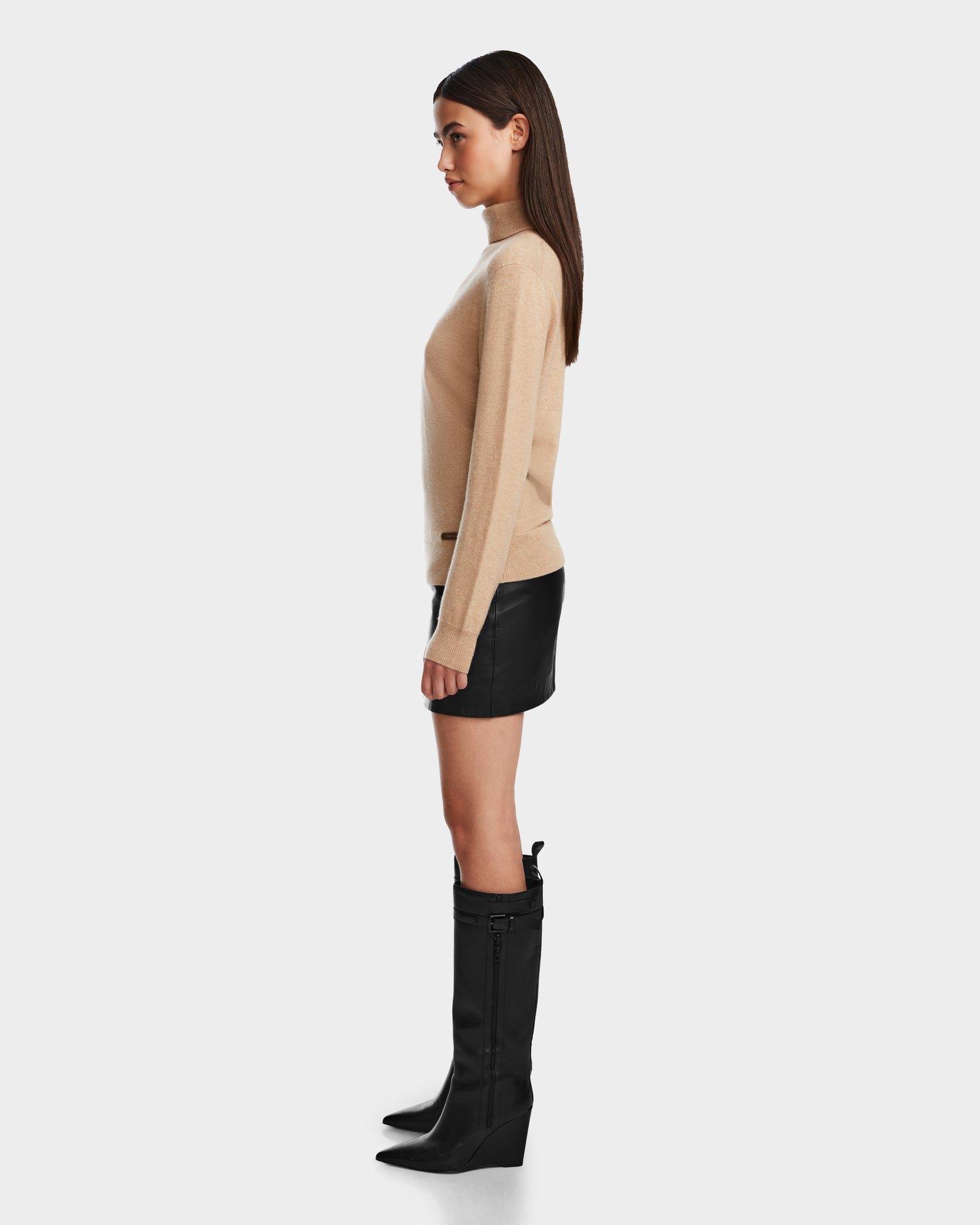 A woman with straight hair and a neutral expression stands in profile against a white background, wearing the Rudsak LIZA - 8524598 Camel premium cashmere sweater, paired with a black leather mini skirt and black knee-high boots.