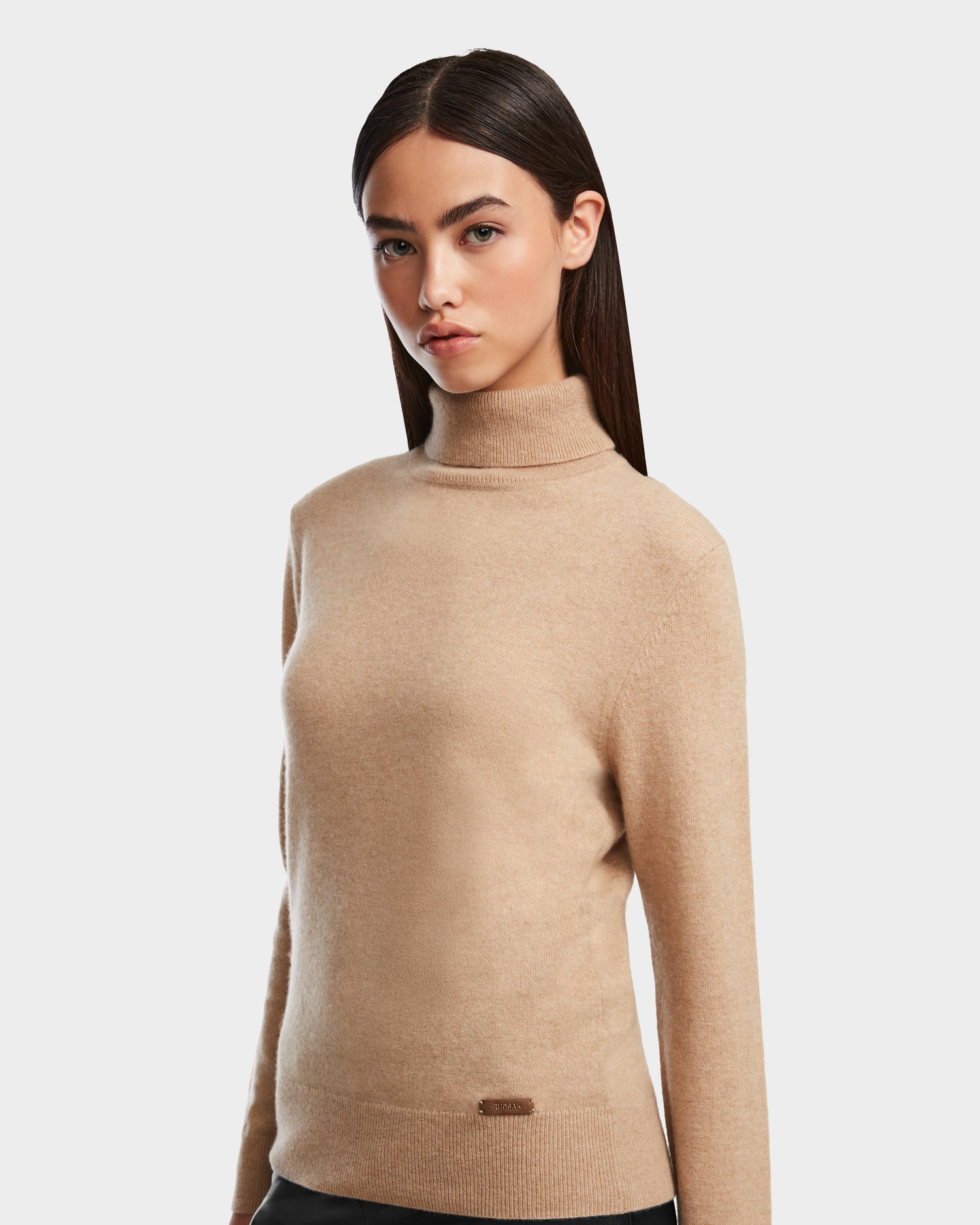 A woman with straight dark hair models the LIZA - 8524598 Camel turtleneck sweater by Rudsak, paired with black pants, standing against a plain light gray background and facing forward with a neutral expression.