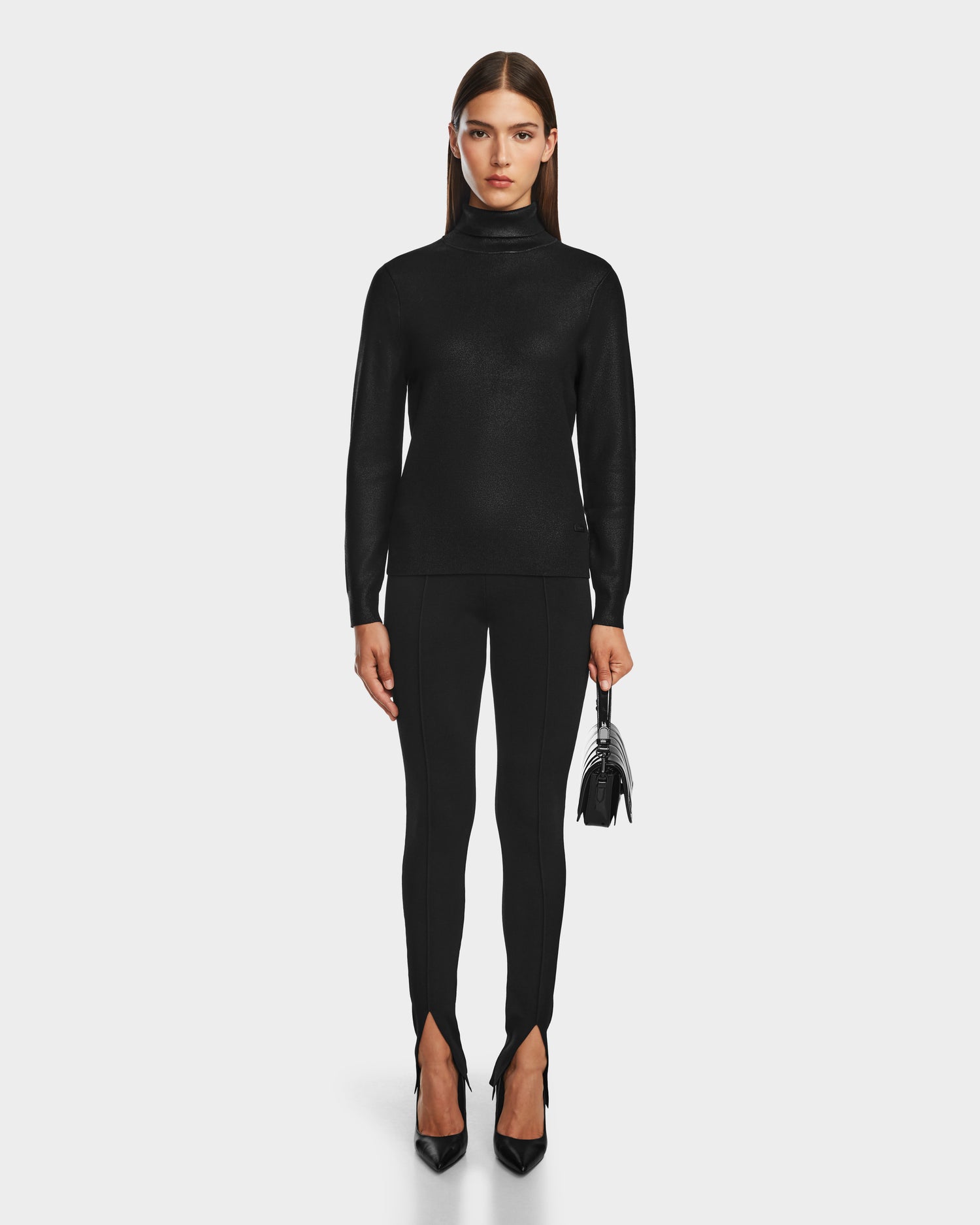 A woman with straight brown hair wears the Rudsak LIZA - 8524598 Metallic Black turtleneck sweater, black fitted pants with front ankle slits, black heels, and holds a small handbag against a plain white background.
