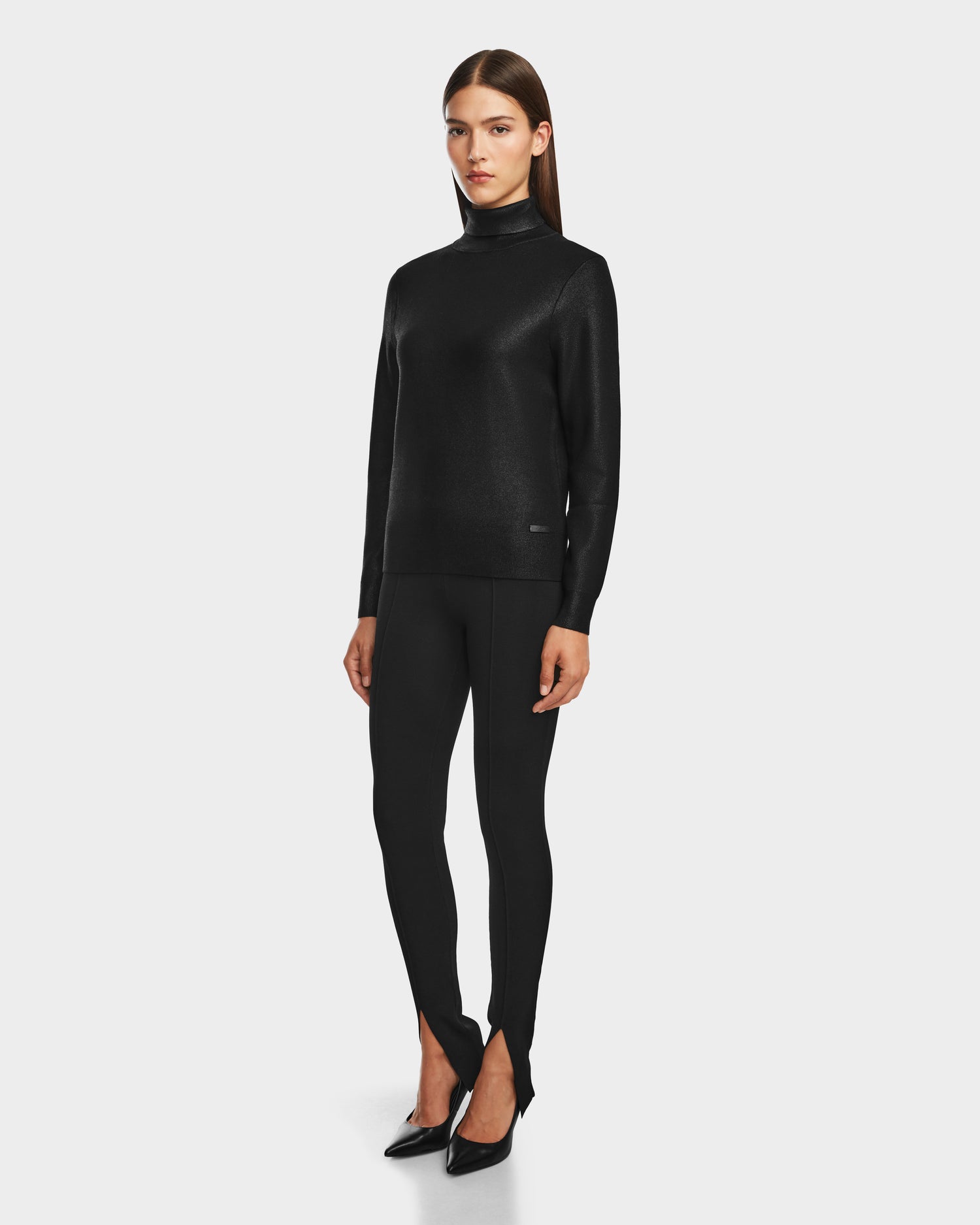 A woman faces forward wearing the Rudsak LIZA - 8524598 Metallic Black turtleneck sweater, paired with black fitted pants with front ankle slits and black pointed-toe heels, set against a plain white background.