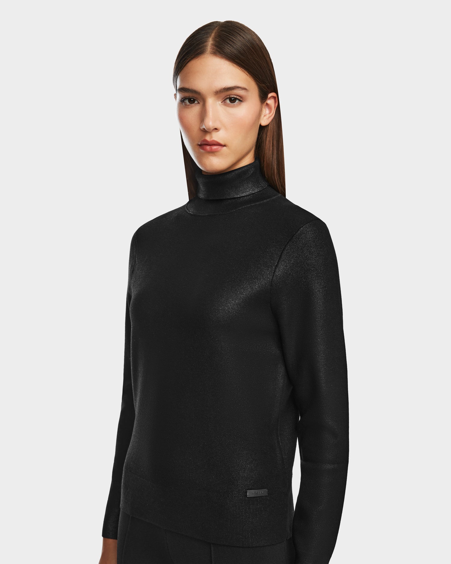 A woman with straight brown hair wears the Rudsak LIZA - 8524598 Metallic Black fitted, long-sleeve women's turtleneck against a plain light gray background, looking slightly to the side with a neutral expression.