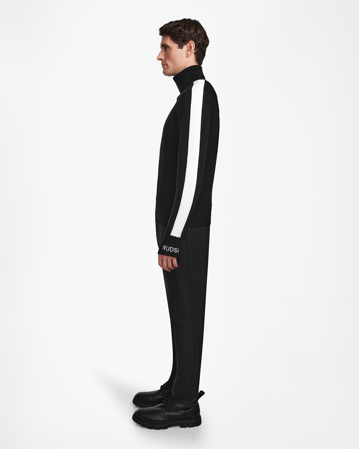 A man stands in profile against a white background, wearing the Rudsak LUKAS - 6524543 Black mens knit turtleneck with a white sleeve stripe, black pants and boots. RUDSAK is visible on his sleeve.