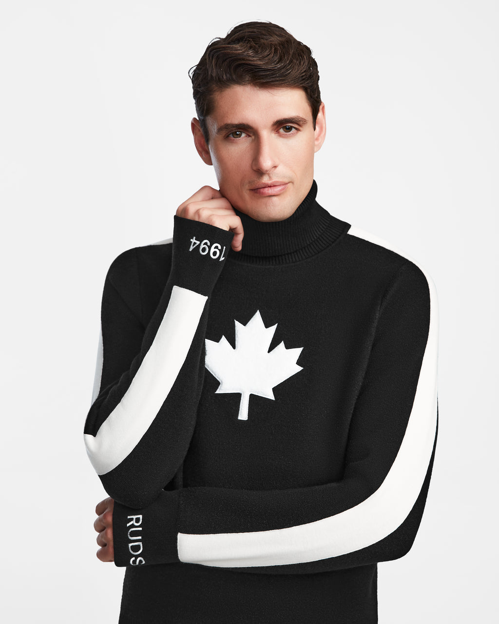 A man wears the Rudsak LUKAS - 6524543 Black men’s knit turtleneck sweater with white sleeve stripes and a maple leaf emblem, posing thoughtfully against a plain white background—embodying winter sports style.