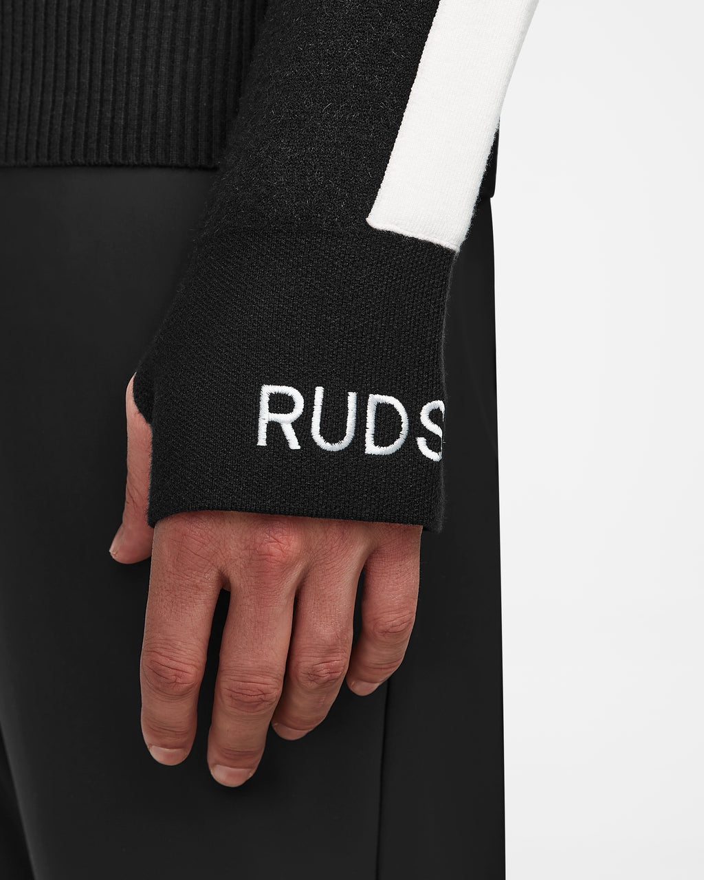 A close-up of a person’s left hand wearing the Rudsak LUKAS - 6524543 Black men’s knit turtleneck sweater with a white vertical stripe and partial RUDS embroidery near the cuff against a minimalistic background.