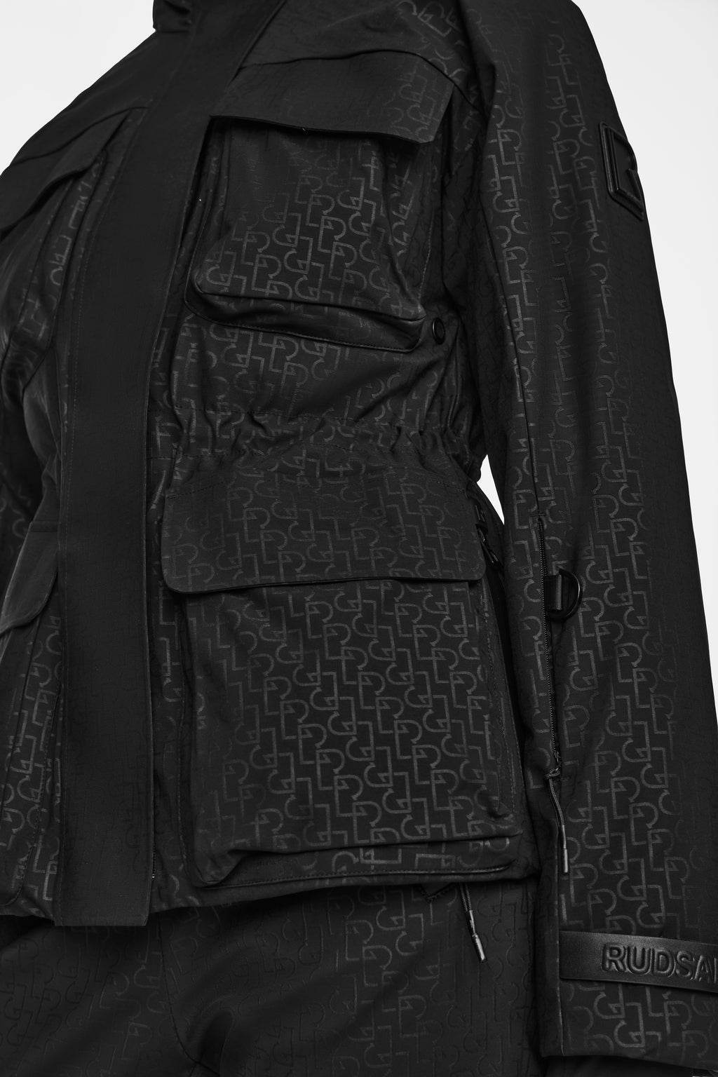 Close-up of the Rudsak LUX MO - 8124570 Monogram Print Black ski jacket and pants set, featuring large flap pockets, a waist drawstring, RECCO rescue technology, and a Rudsak patch on the sleeve.
