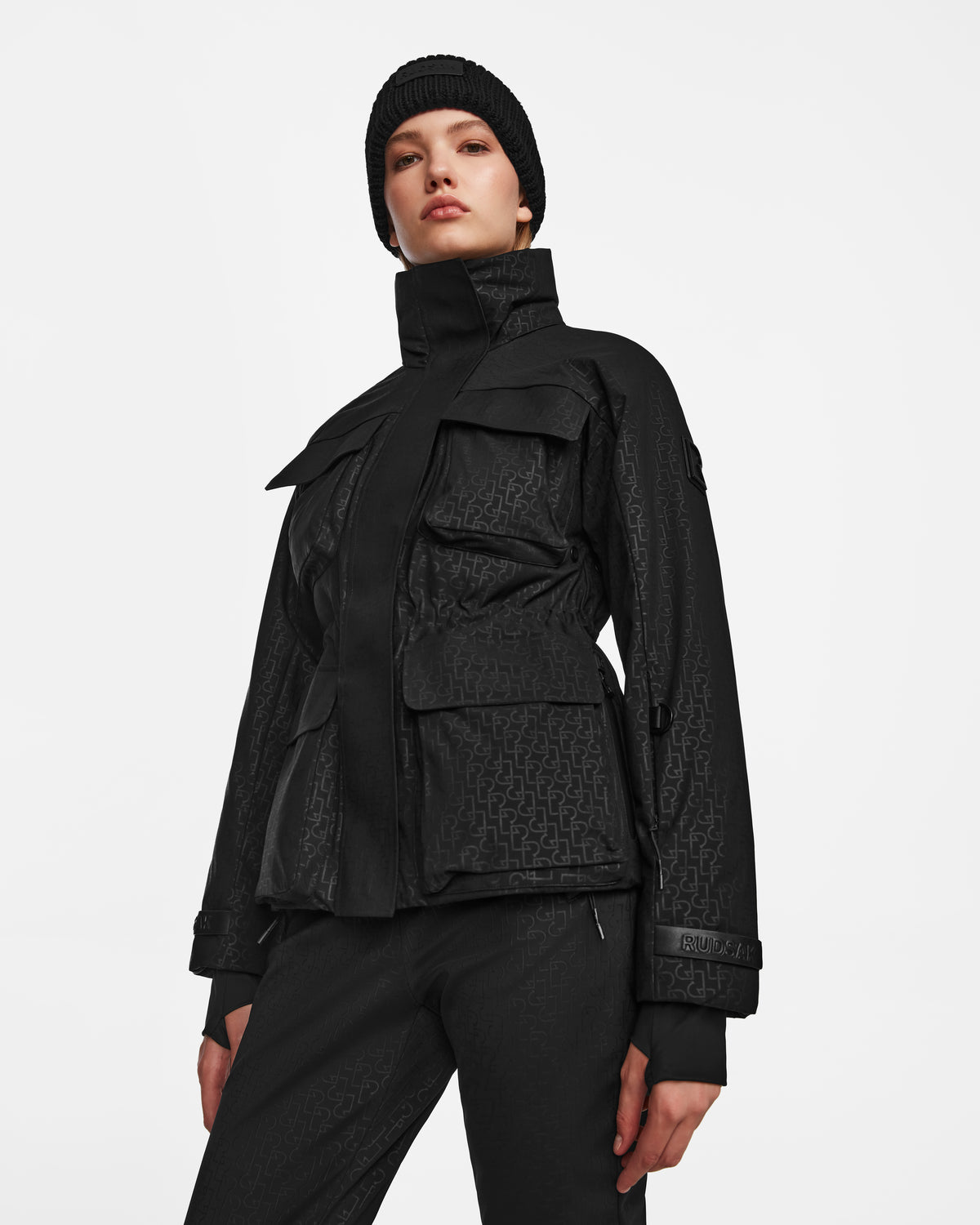 A person models Rudsak’s LUX MO - 8124570 Monogram Print Black ski jacket with RECCO technology and multiple pockets, paired with matching black pants and a black beanie, posing confidently against a white background.