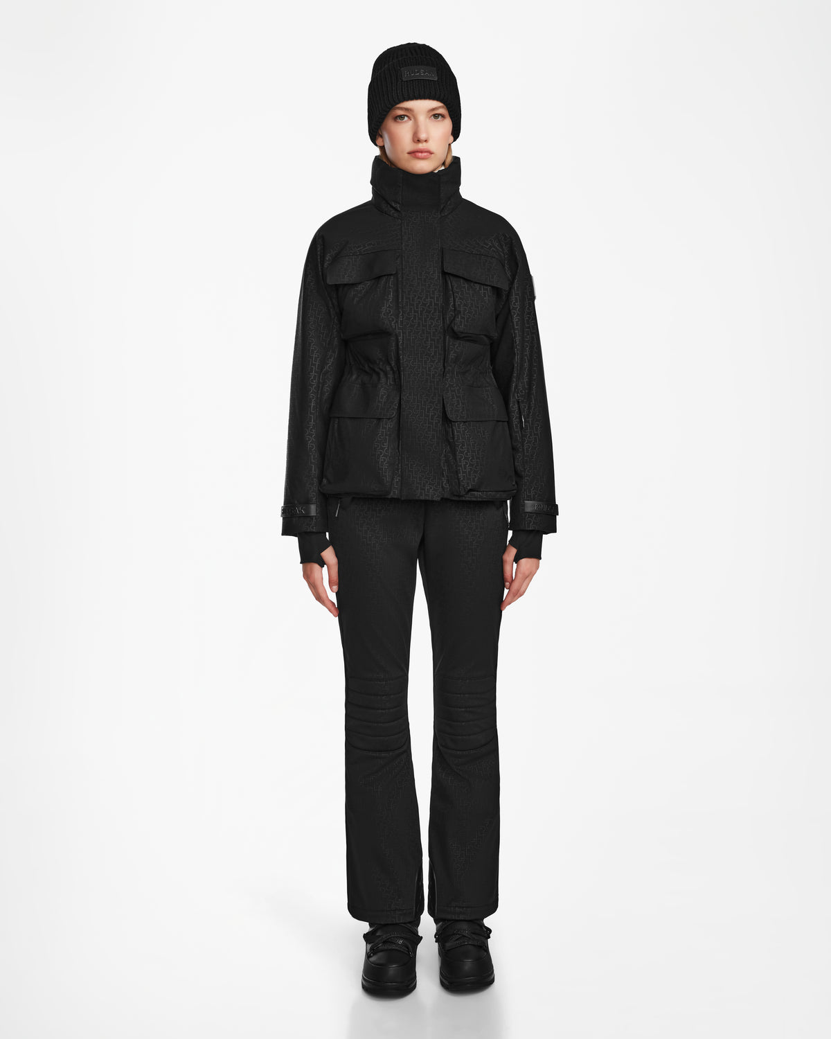 A person stands facing forward against a white background, wearing a Rudsak LUX MO - 8124570 Monogram Print Black ski jacket with large pockets, paired with black pants, shoes, and a black beanie for a sleek monochromatic look.