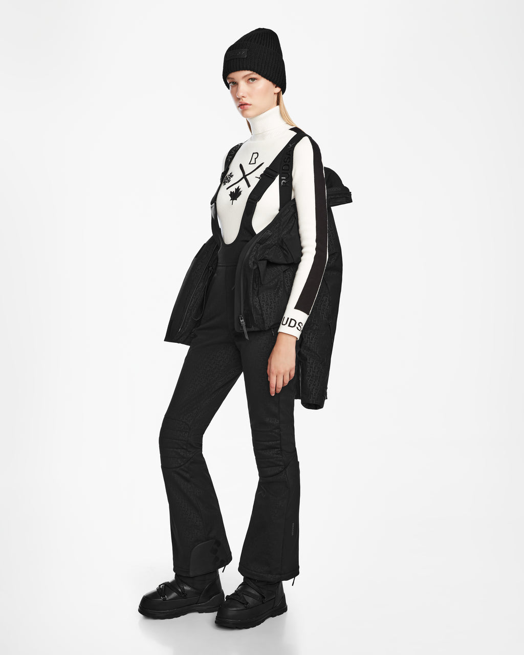 A woman stands against a white background wearing Rudsaks LUX MO - 8124570 Monogram Print Black ski jacket draped over her shoulders, styled with a black beanie, graphic white turtleneck, black overalls, and chunky black boots.