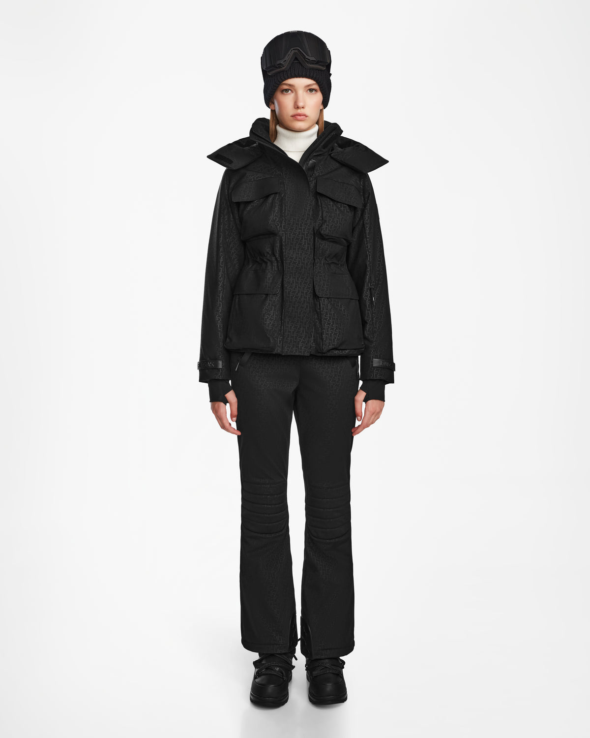 A woman models the Rudsak LUX MO - 8124570 Monogram Print Black ski jacket and matching pants against a white background, paired with black shoes, a white turtleneck, and a black cap. She gazes at the camera with a neutral expression.