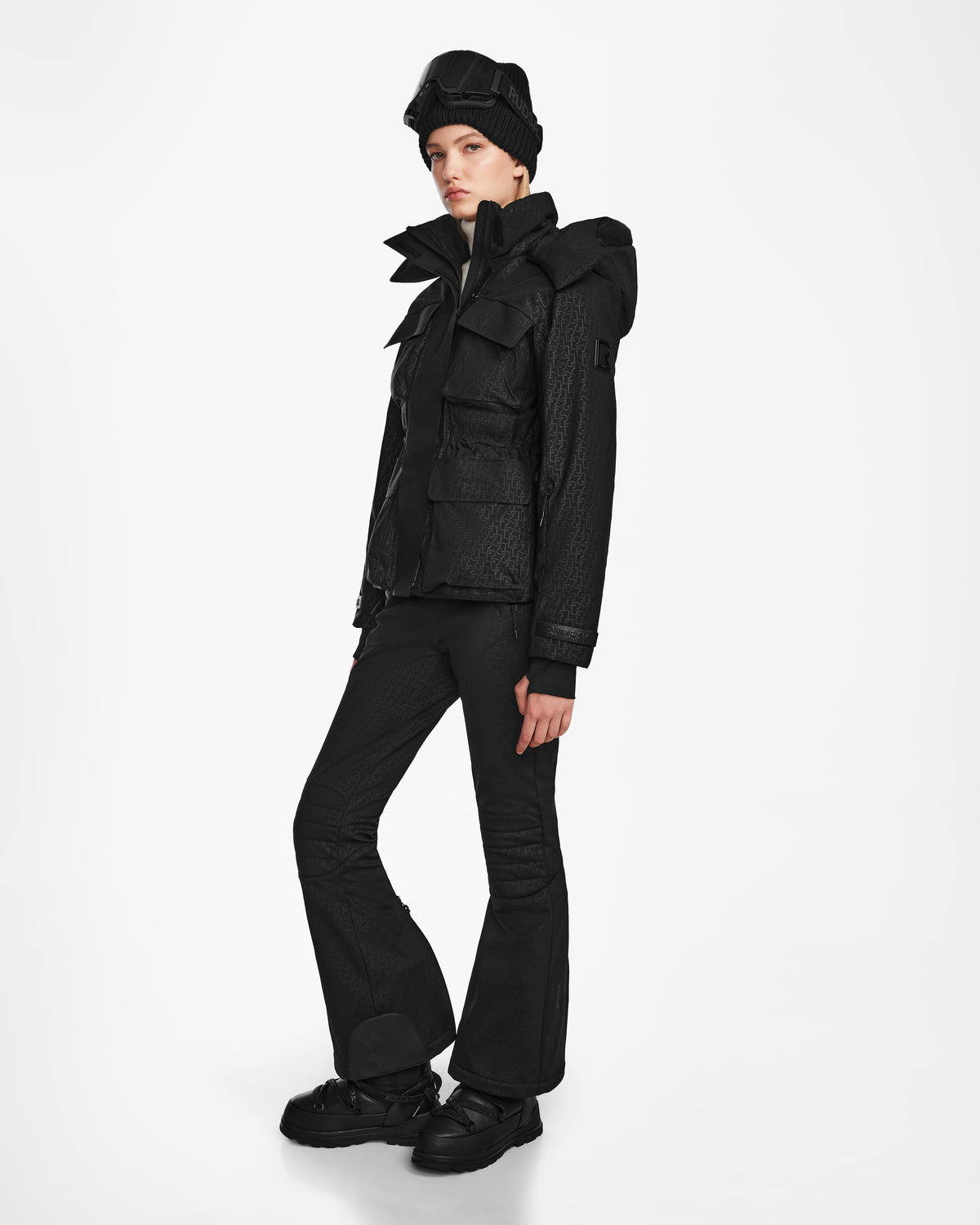 A person stands against a white background dressed in a Rudsak LUX MO - 8124570 Monogram Print Black ski jacket with large pockets, flared pants, chunky boots, a knit beanie, and ski goggles.