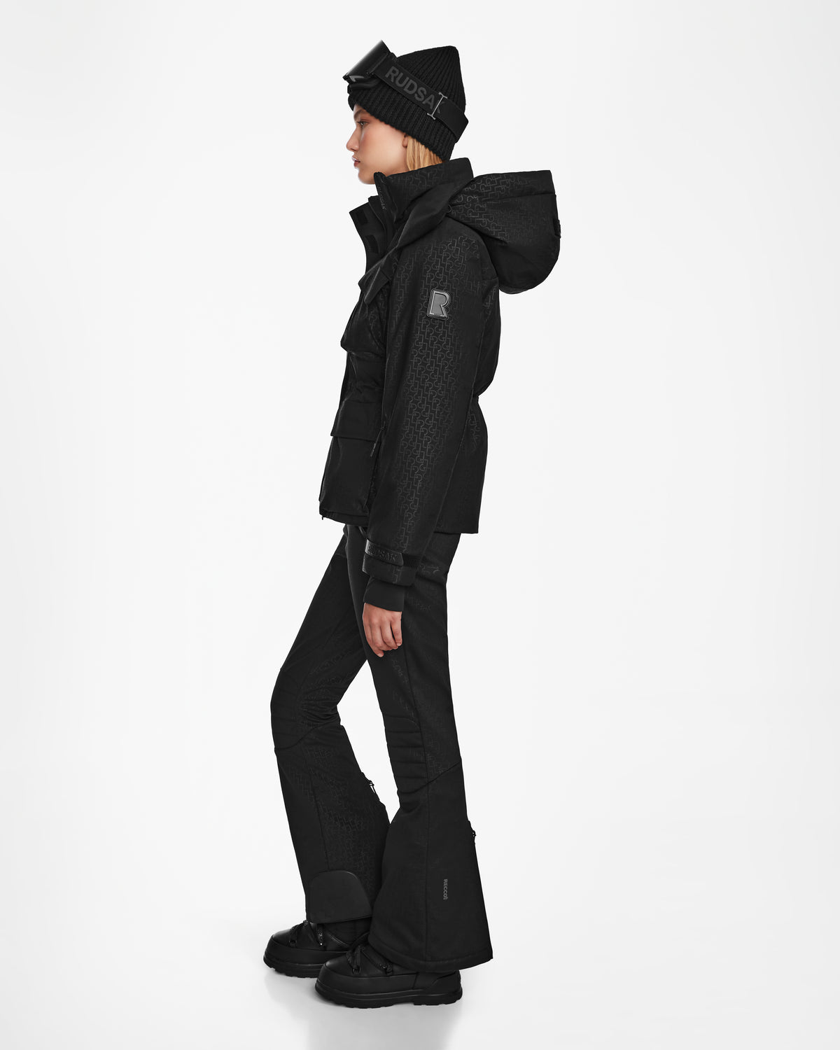 A person in profile wears the Rudsak LUX MO - 8124570 Monogram Print Black women’s ski jacket with a hood, matching pants, boots, and a beanie. The plain white background highlights its RECCO rescue technology for cold weather.