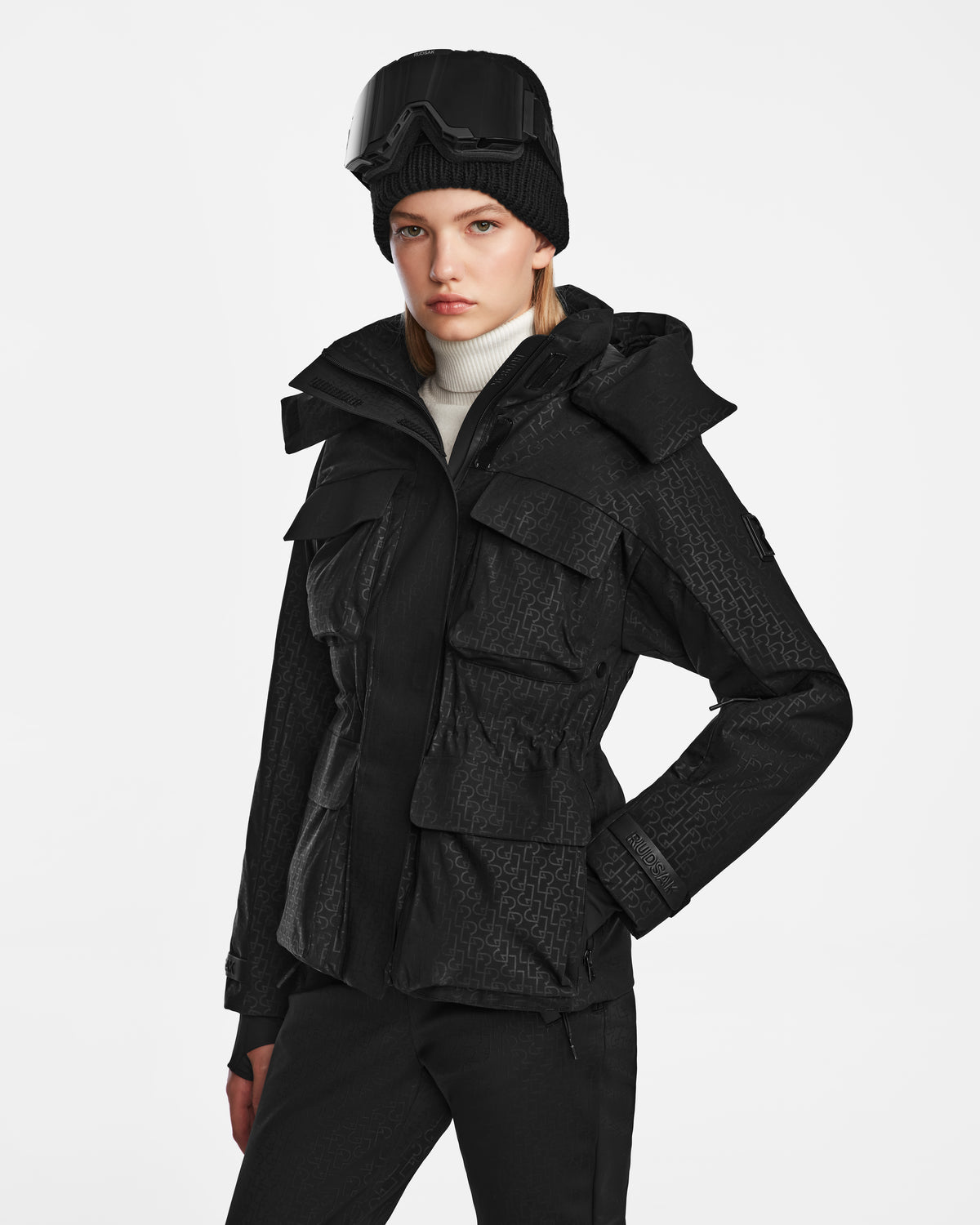 A person wearing the Rudsak LUX MO - 8124570 Monogram Print Black waterproof ski jacket stands against a plain white background, styled with black pants, a black knit beanie, and black ski goggles.