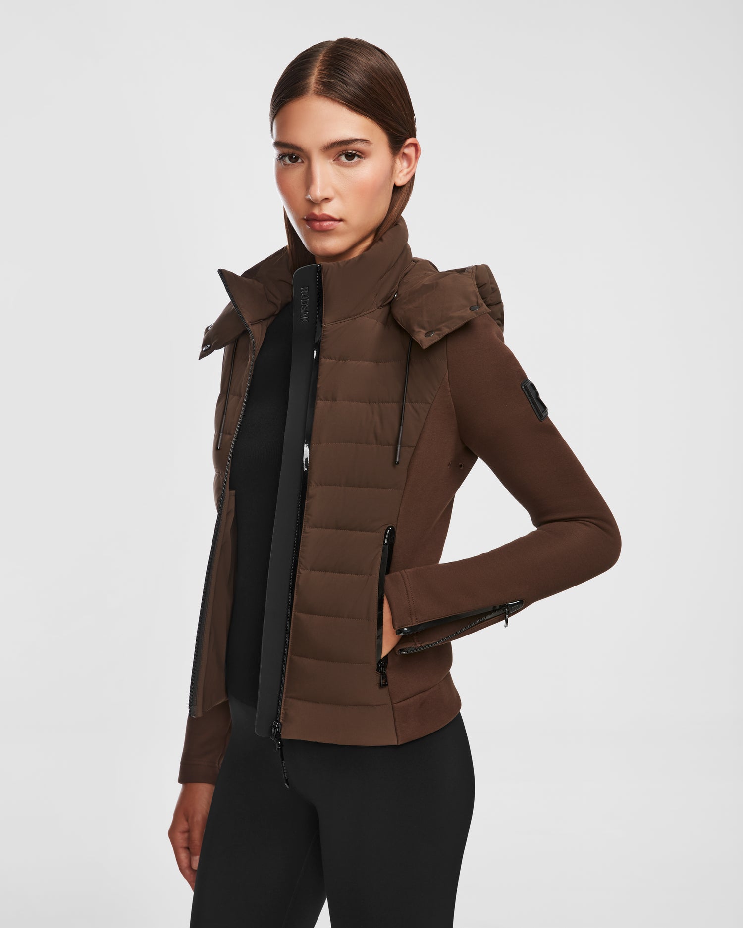 A woman with straight brown hair models the Rudsak MABS - 8124825 Espresso, a fitted lightweight short jacket with a high collar, zipper pockets, and detachable hood, posing confidently against a plain white background.