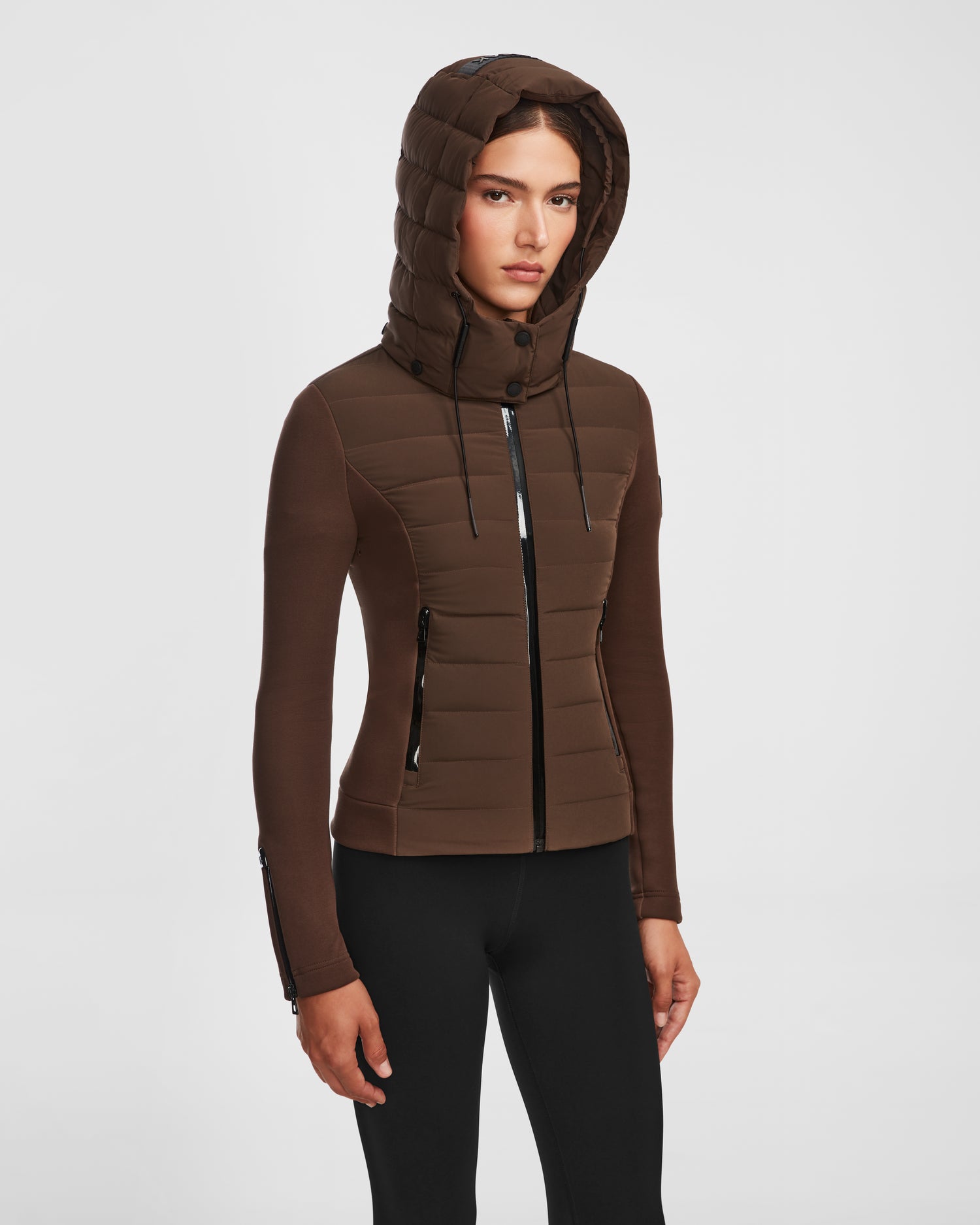 A woman wearing the Rudsak MABS - 8124825 Espresso fitted womens jacket with a hood and black zipper details, paired with black pants, stands against a plain white background.
