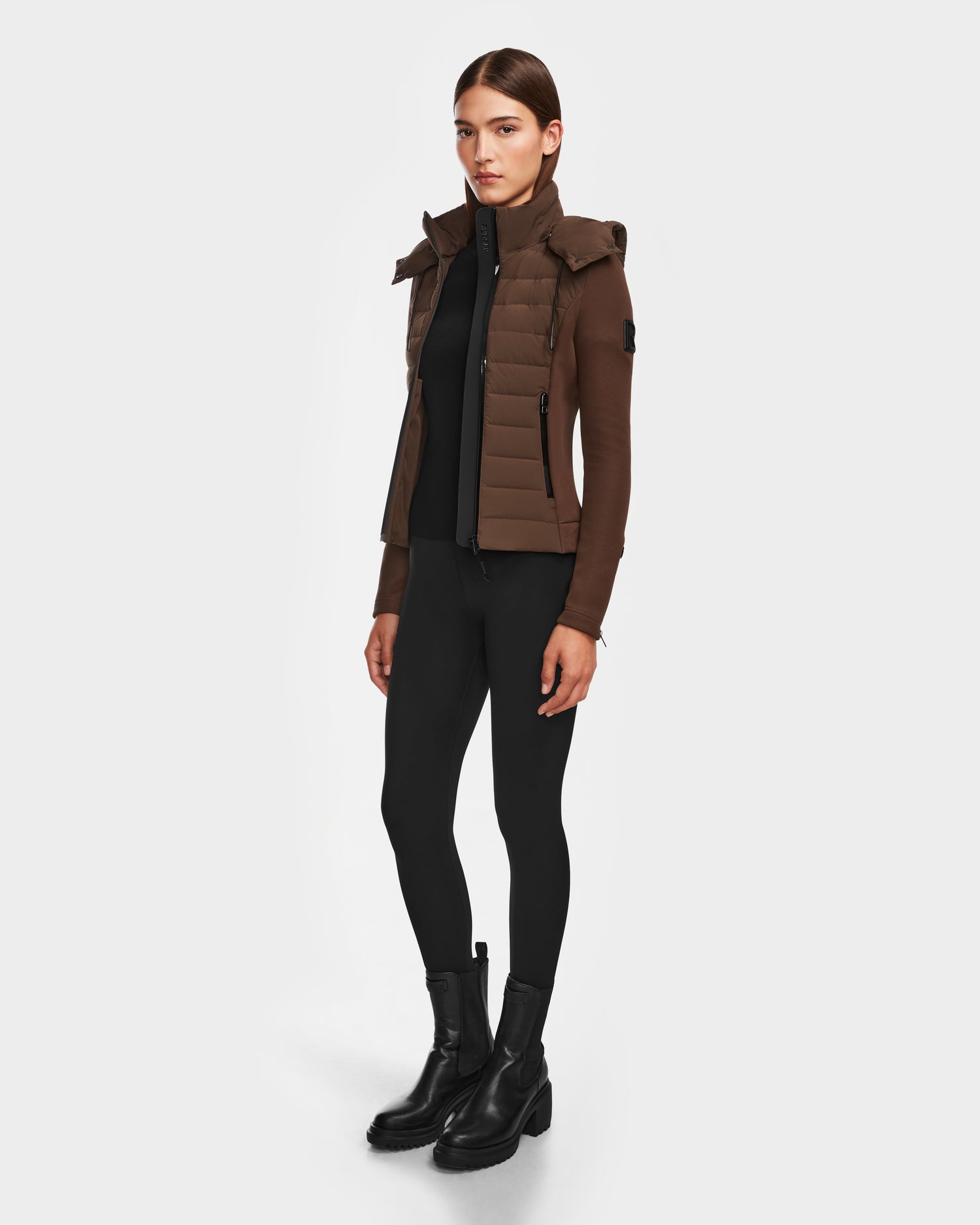 A woman with straight brown hair and a neutral expression poses against a white background wearing the Rudsak MABS - 8124825 Espresso women’s jacket with hood, paired with black leggings and black ankle boots.