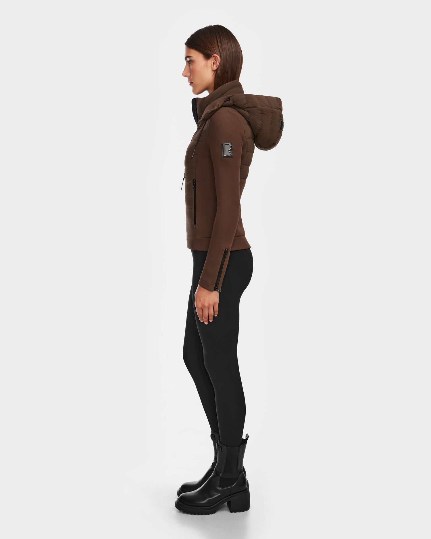 A woman in profile against a white background wears the Rudsak MABS - 8124825 Espresso lightweight brown hooded jacket, paired with black leggings and black boots.