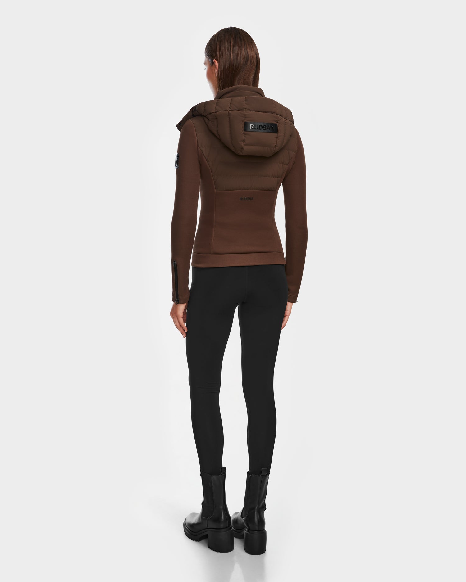 A woman faces away, wearing the Rudsak MABS - 8124825 Espresso fitted brown neoprene jacket with a hood, paired with black leggings and boots against a plain white background.