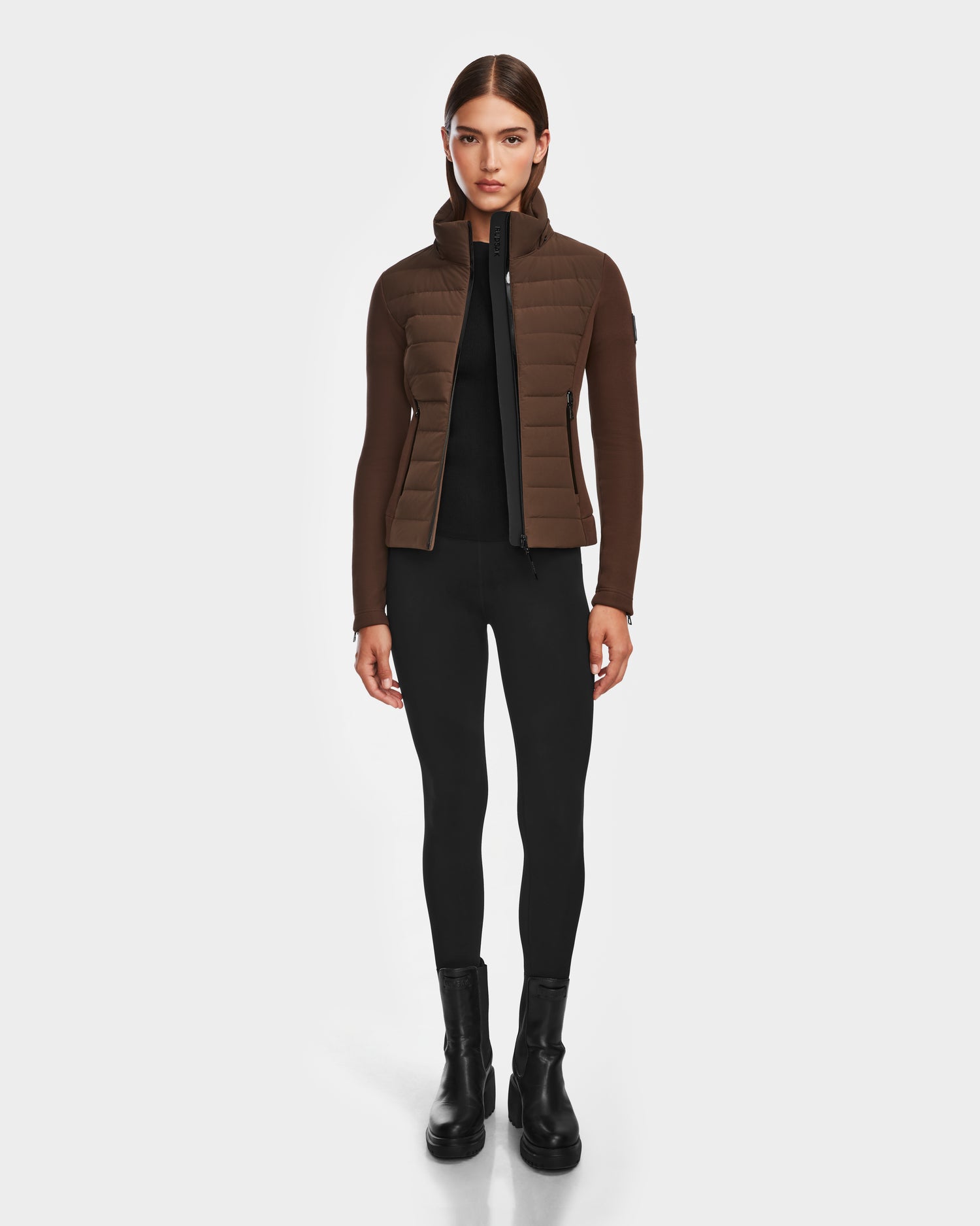 A woman faces forward against a white background, wearing the Rudsak MABS - 8124825 Espresso womens jacket over a black top, black leggings, and black ankle boots.