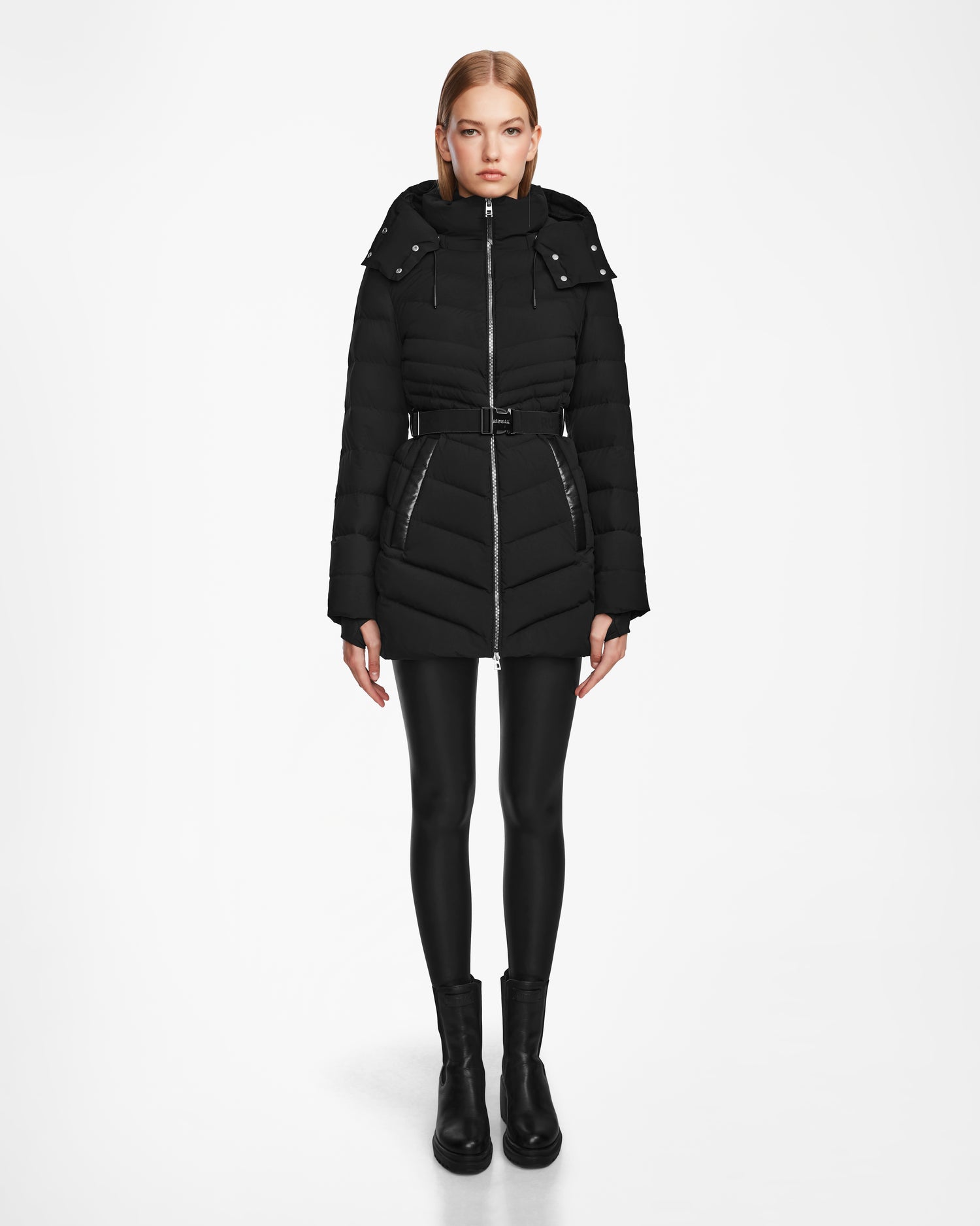 A woman faces forward against a white background wearing the Rudsak MALAYA M - L123554 Black women’s down puffer jacket with a hood, belted waist, and herringbone quilting, paired with black leggings and ankle boots.