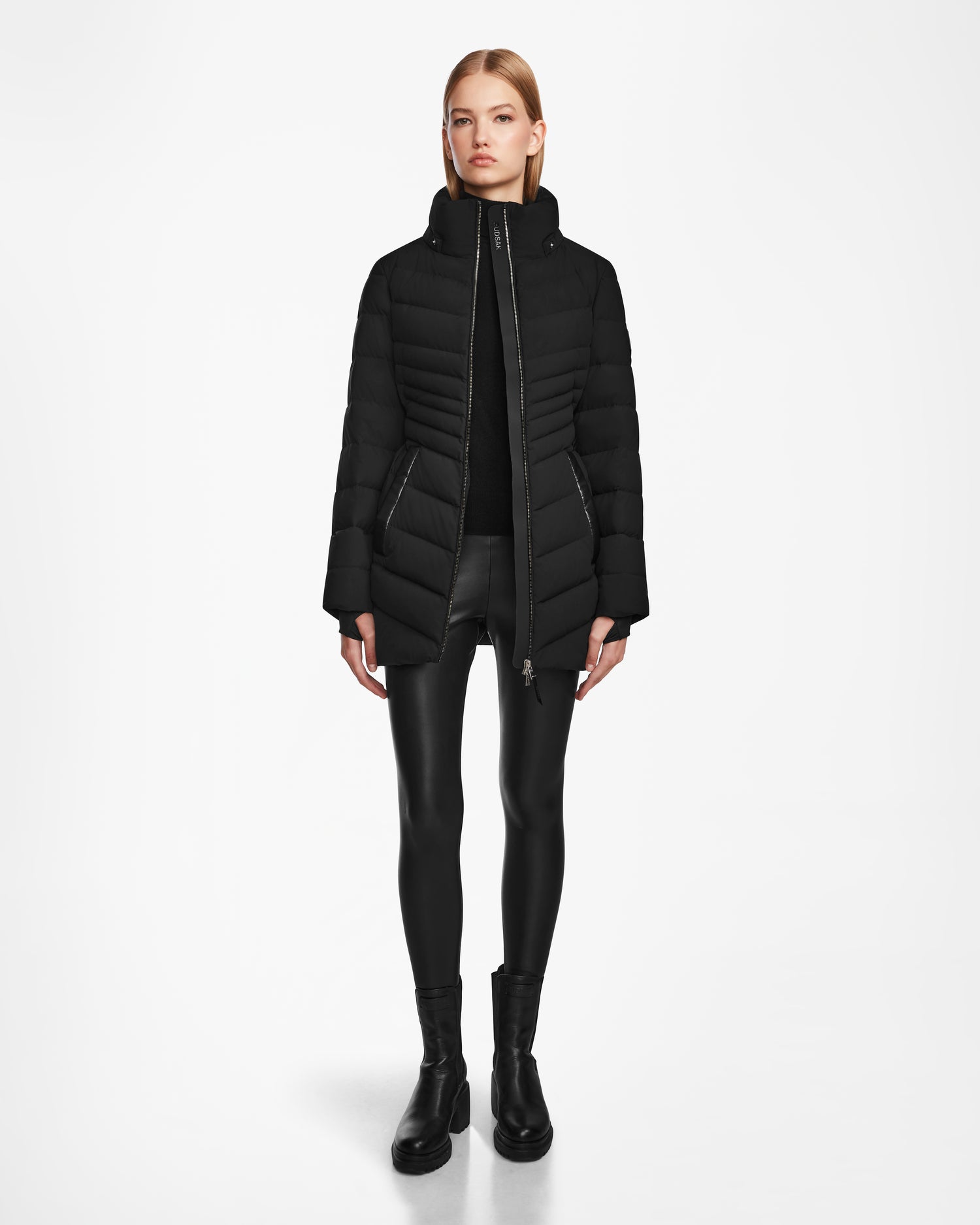 A woman faces forward against a white background, wearing the Rudsak MALAYA M - L123554 Black womens down puffer jacket with a sleek herringbone quilting, paired with a black top, leather-look leggings, and ankle boots.